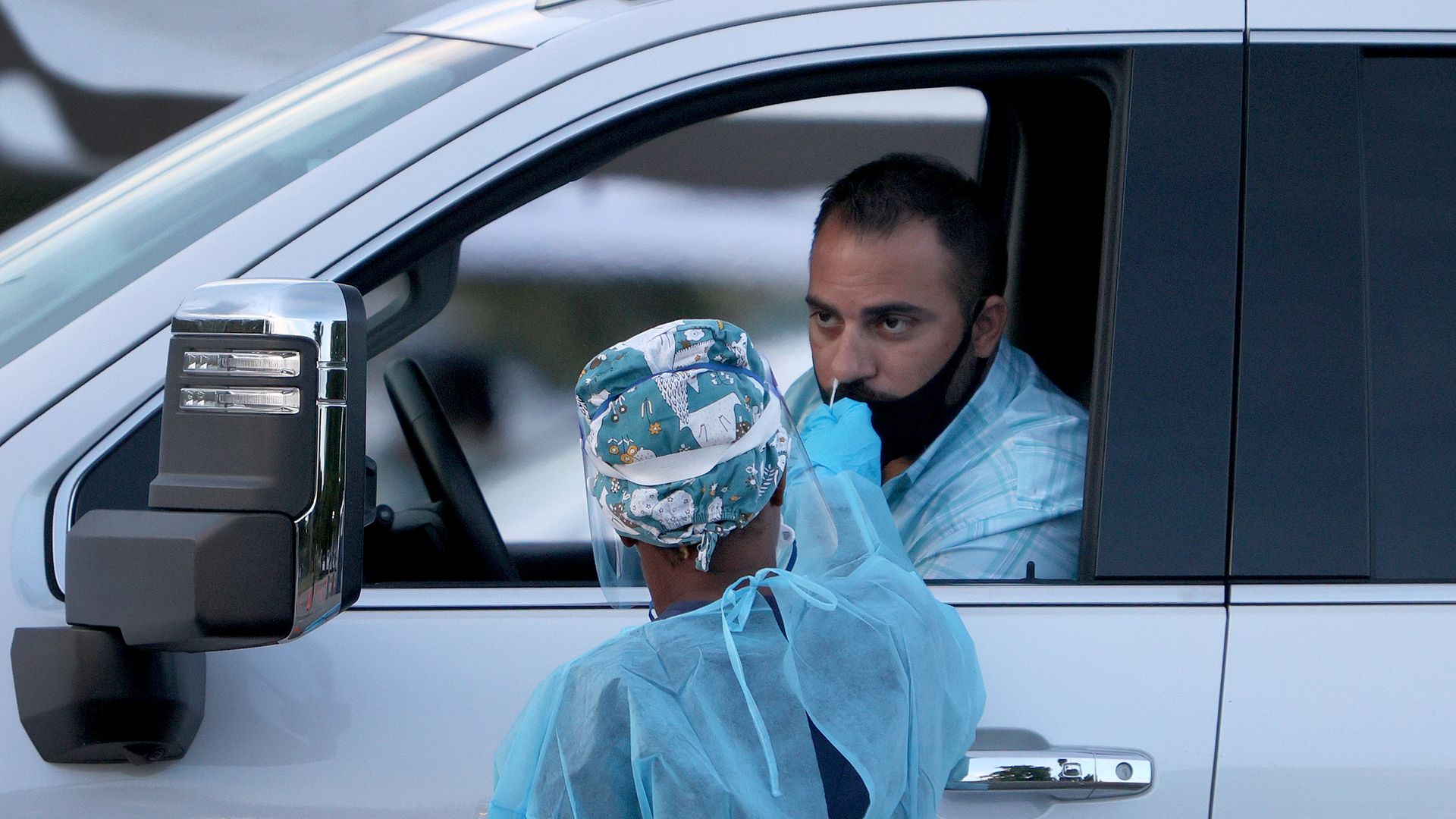 A health care worker in a blue gown gets a COVID test swab from a man in a car.