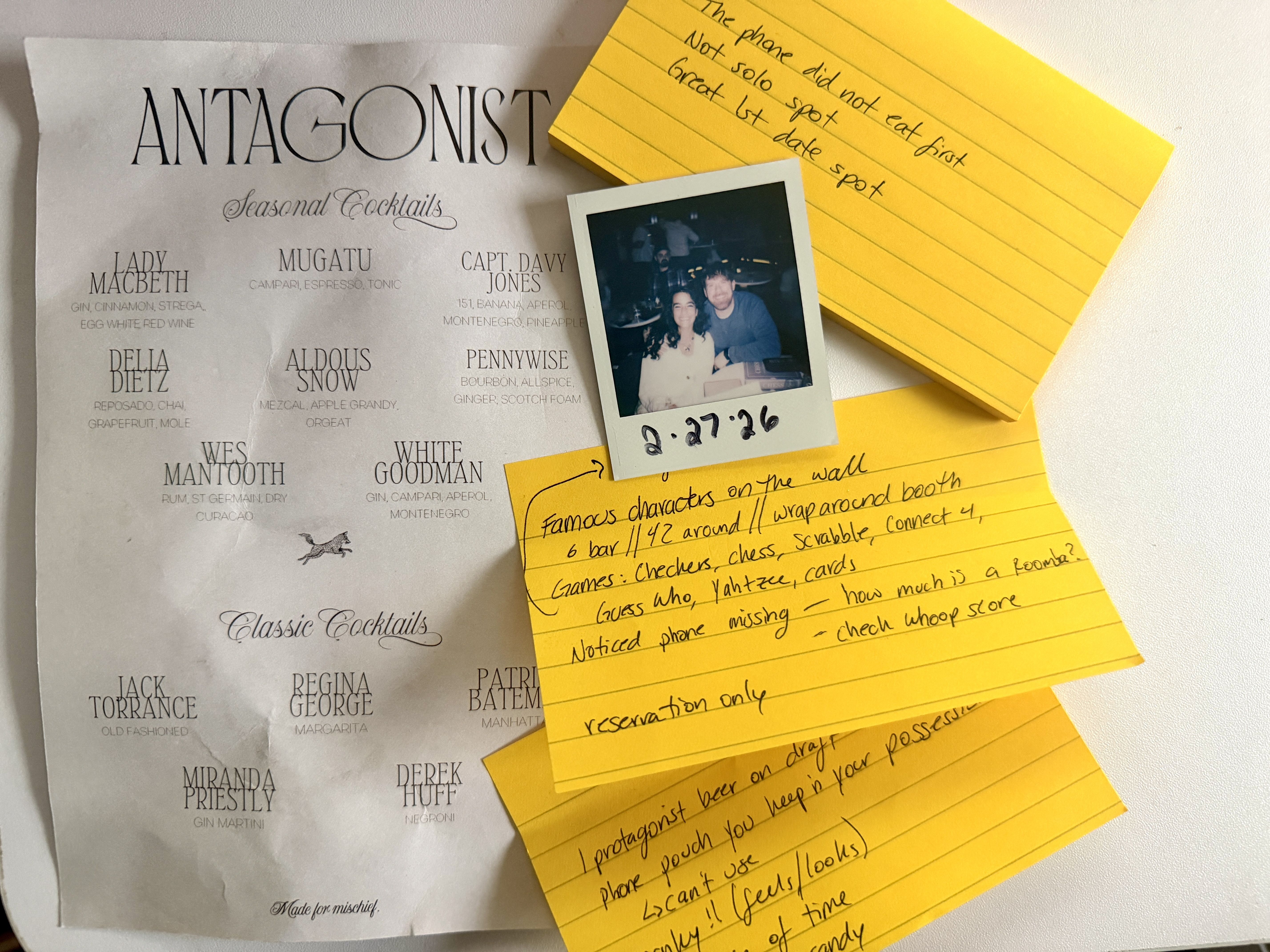 Cocktail menu titled "ANTAGONIST" with seasonal and classic drinks named after characters. Yellow index cards with handwritten notes and a dated Polaroid photo of a couple on a table.