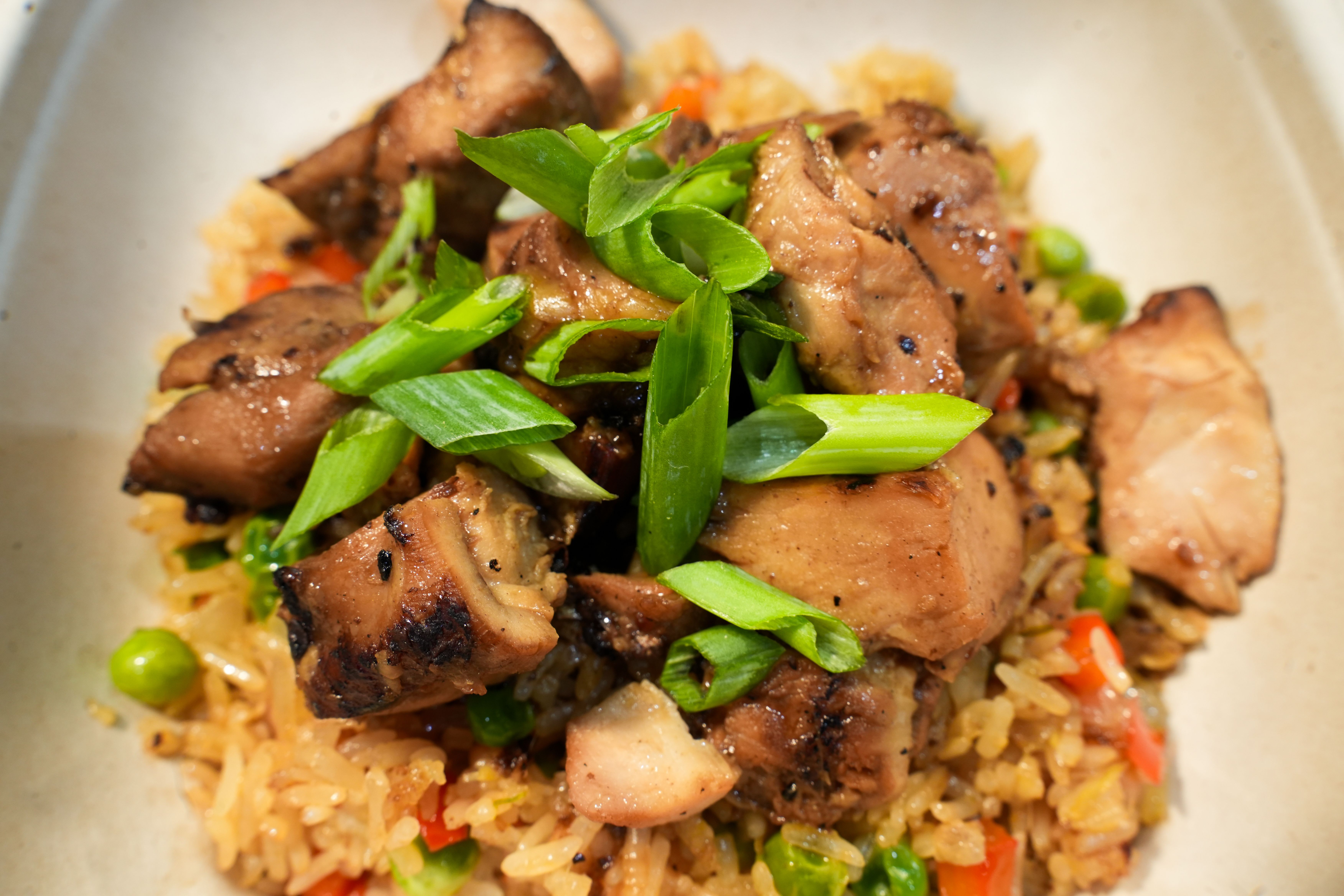 Close-up of grilled chicken pieces and chopped green onions atop fried rice with peas and red bell peppers in a beige bowl.