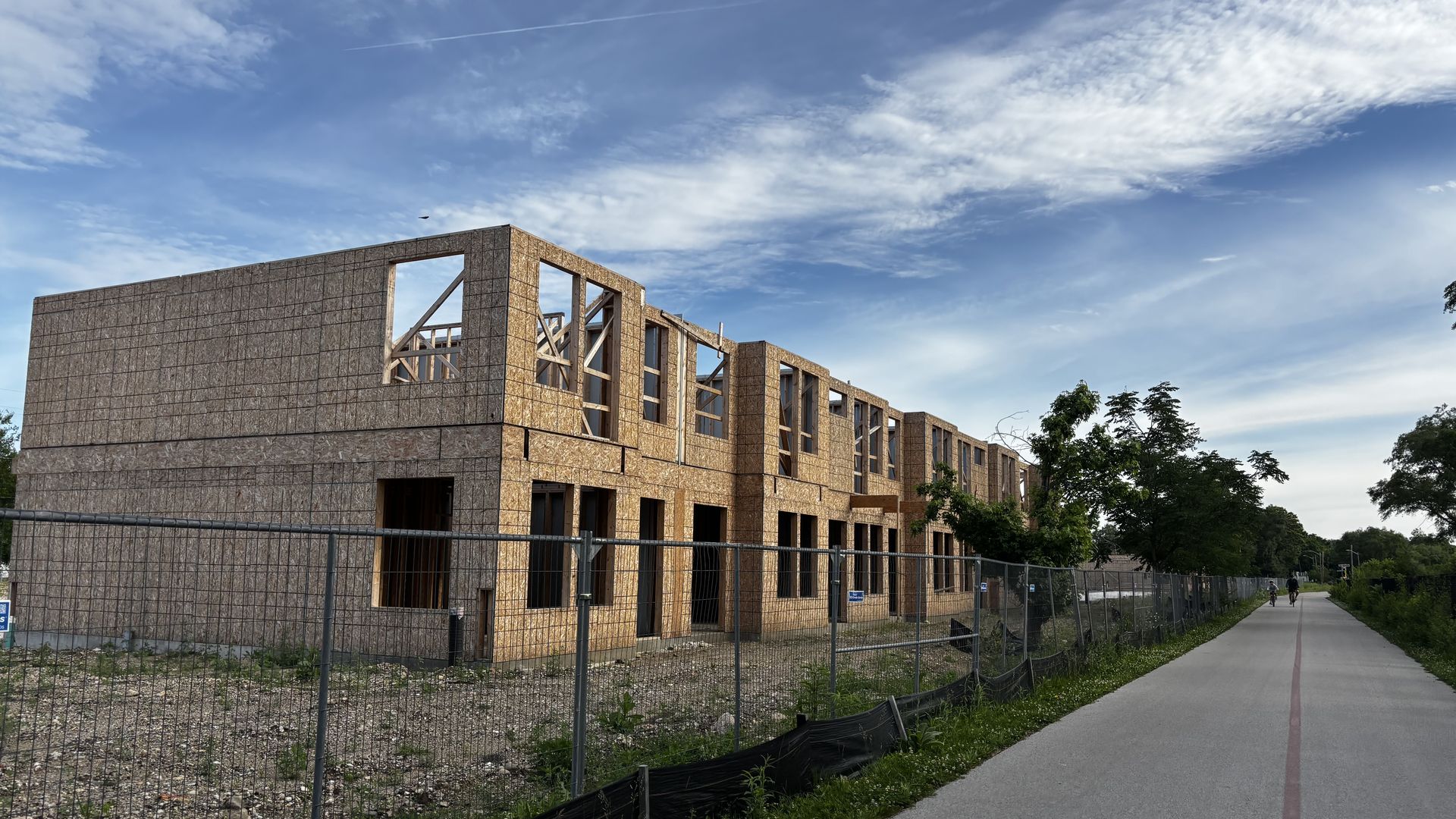 Under-construction, two-story building with plywood sheathing and large window openings; chain-link fence in foreground, a paved path beside, and a blue sky with wispy clouds.