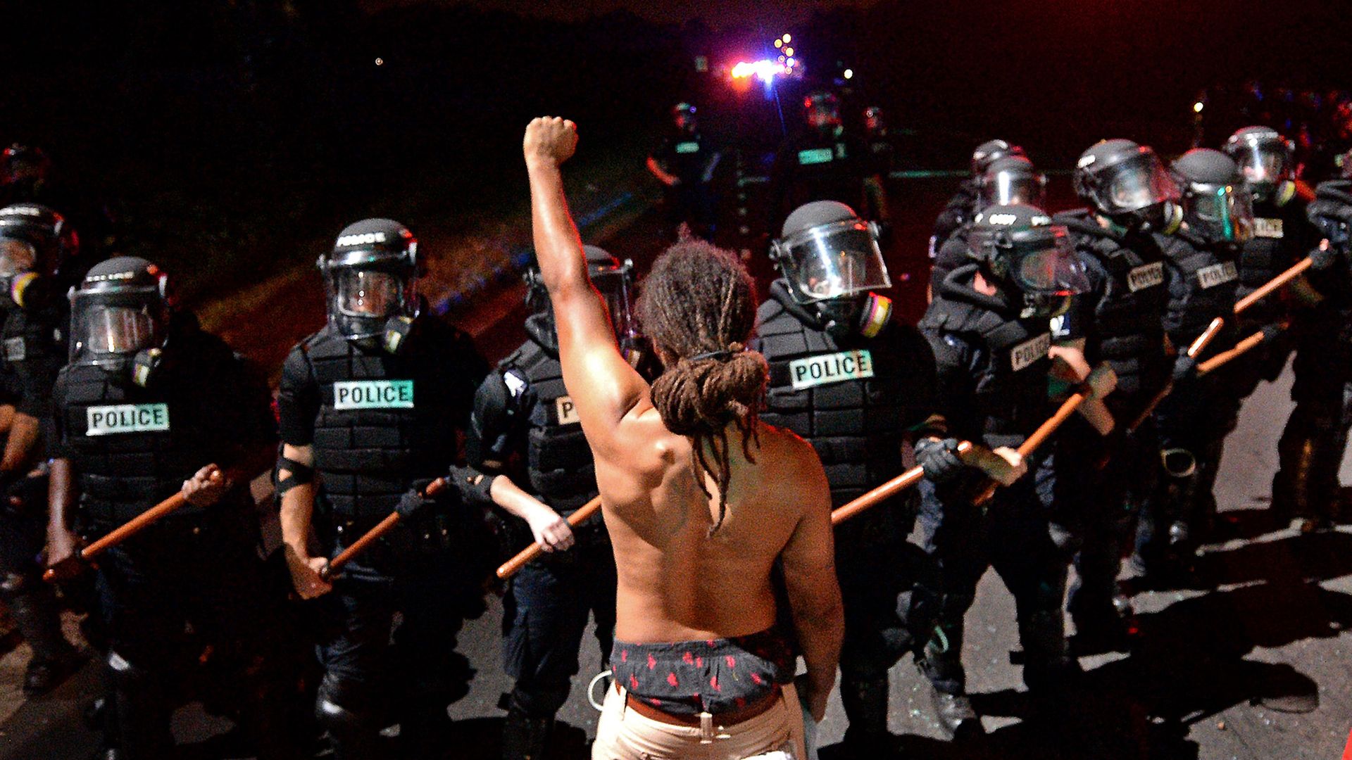 Activist stands in front of line of police, shirtless and holding his fist in the air. 
