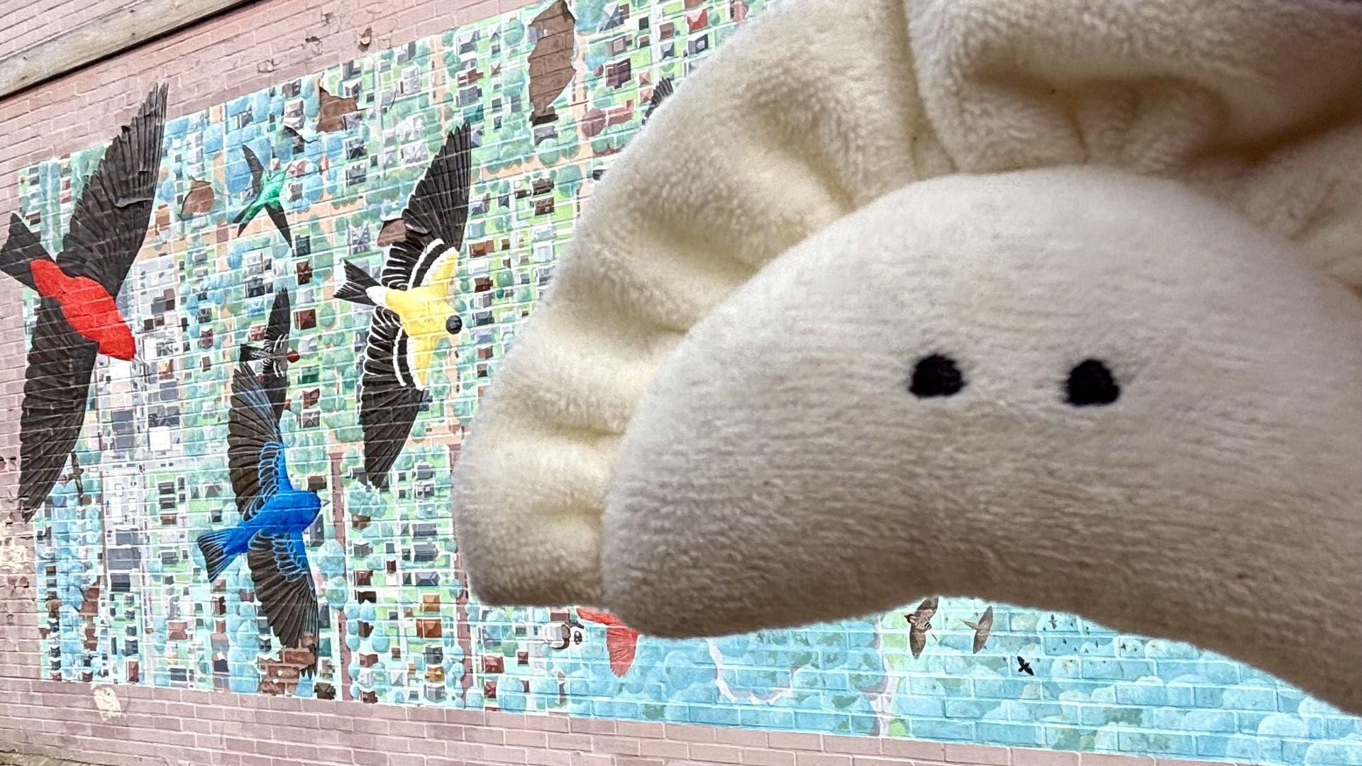 Close-up of a colorful bird mosaic mural on a pink brick wall, with a large beige stuffed animal head partially covering the right side and two black eyes visible.