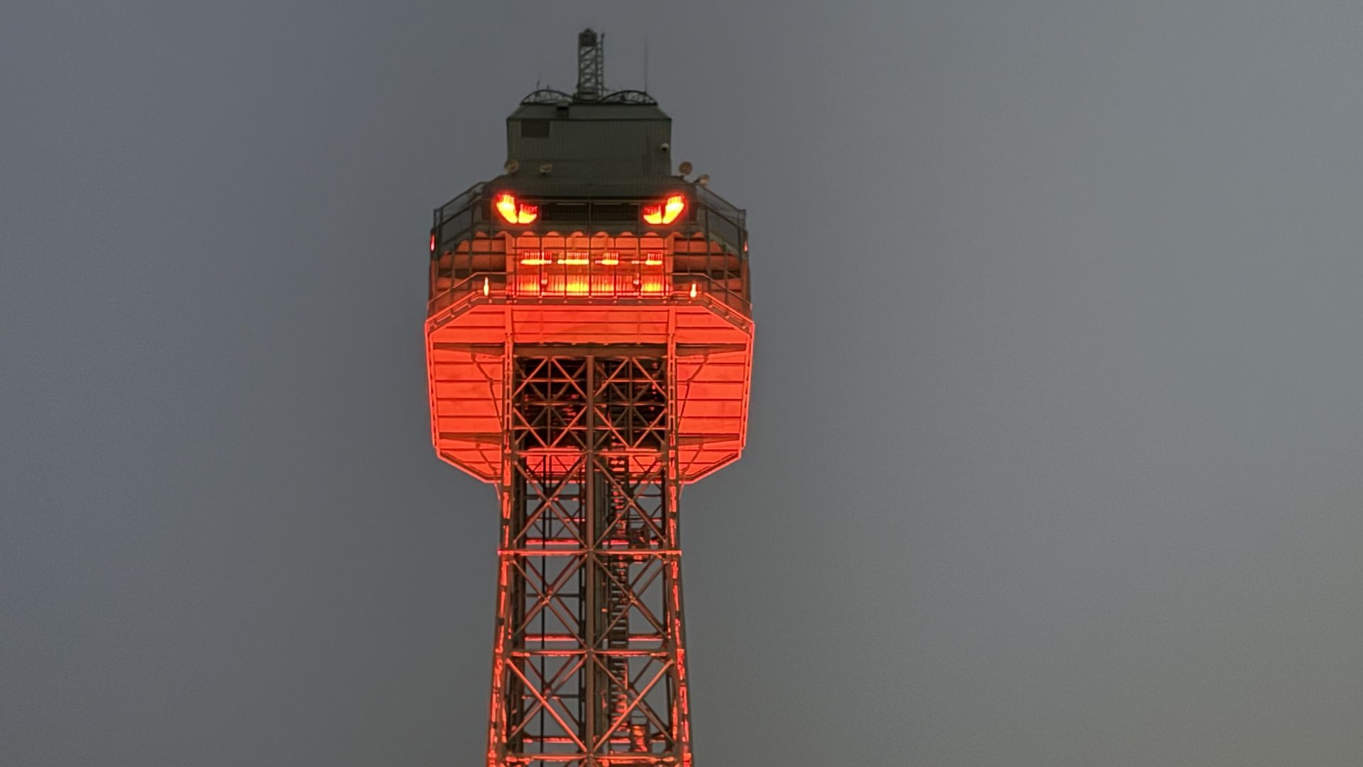 Tall lattice communications tower illuminated in red-orange lights at dusk, with a bright platform near the top and a gray sky behind; trees visible at the base.