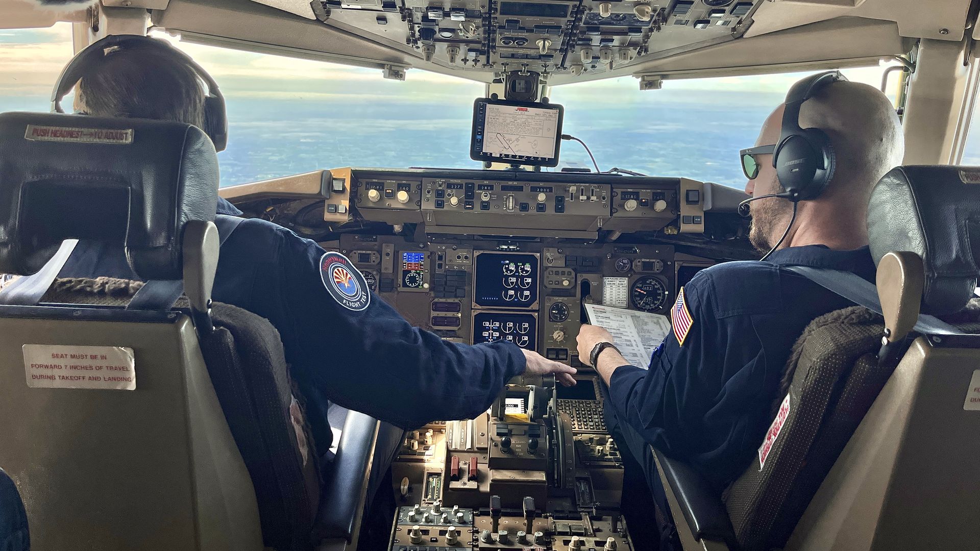 Two pilots wearing headsets and dark uniforms in an airplane cockpit with controls and screens, one holding a checklist, sky visible through the front window.