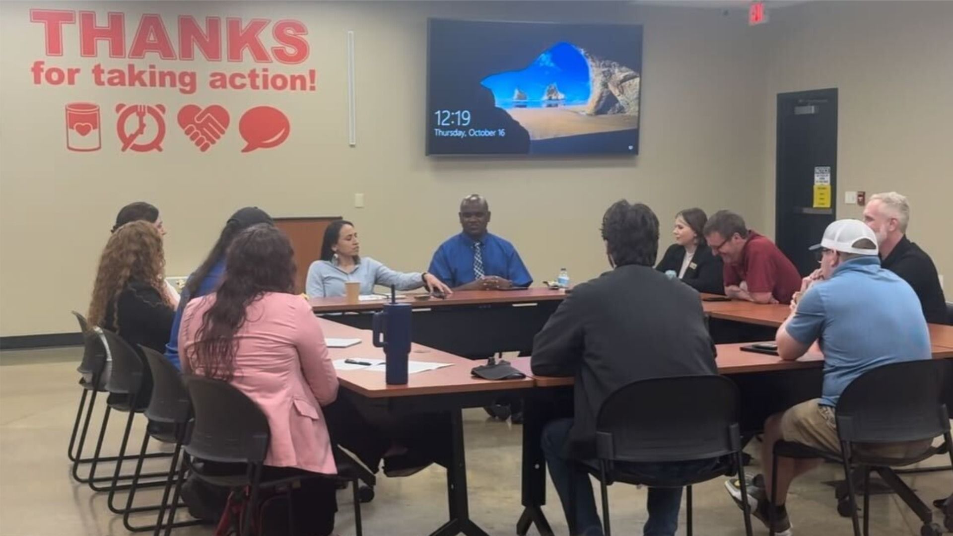 Group of people seated in a meeting room around a U-shaped table with a man in a blue shirt speaking. Wall sign reads "THANKS for taking action!" with icons above.