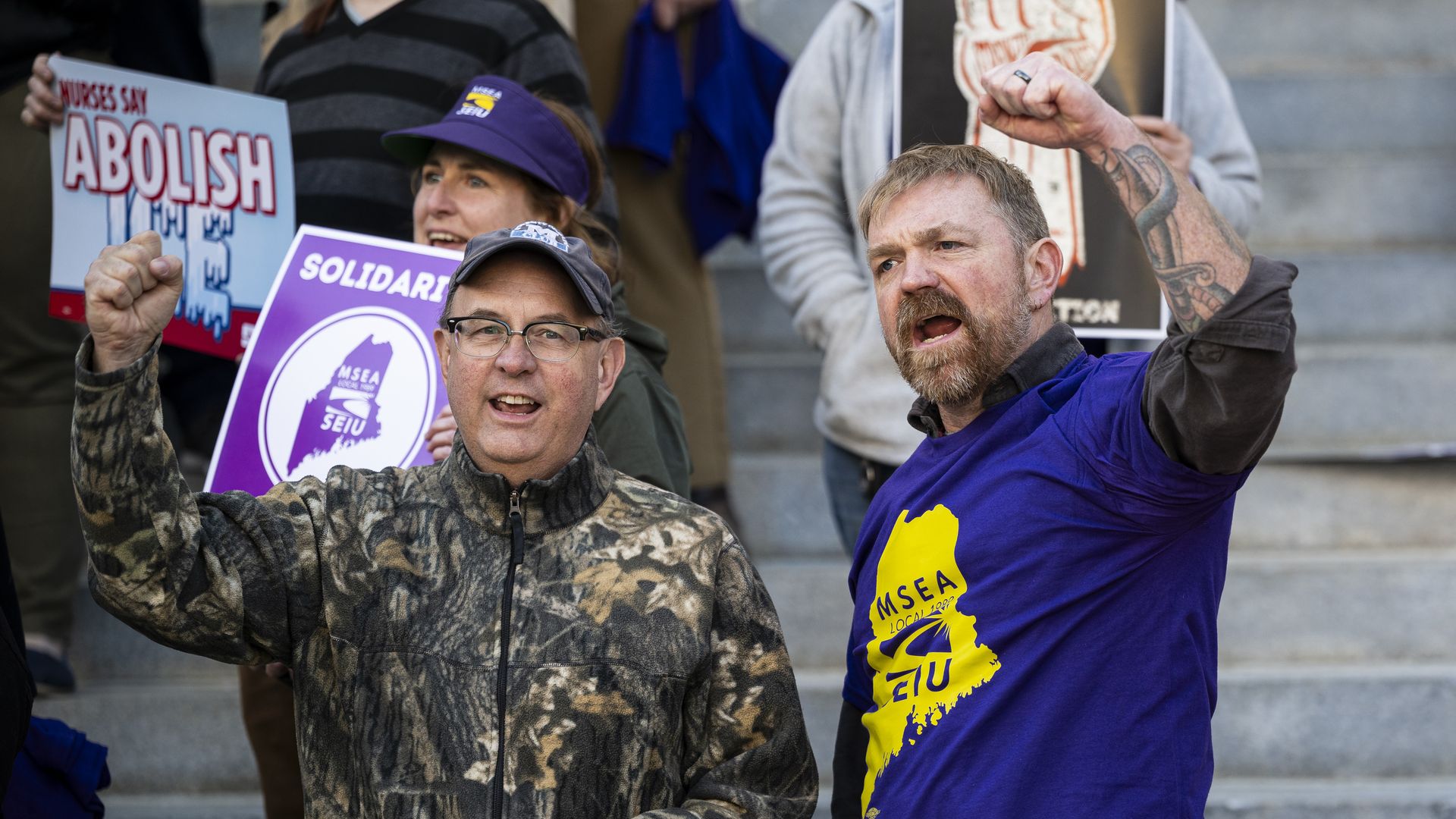 Protesters on concrete steps; central man in a purple shirt raises a clenched fist. Signs read "ABOLISH" and feature a purple emblem; others wear camo and a red blazer, with a woman in the background.