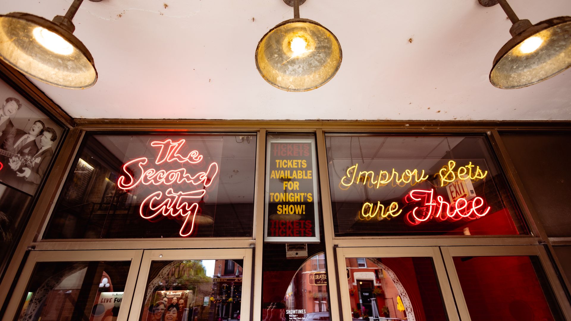 Entrance of The Second City with neon signs in red and yellow saying "The Second City" and "Improv sets are Free" under three rustic ceiling lights.