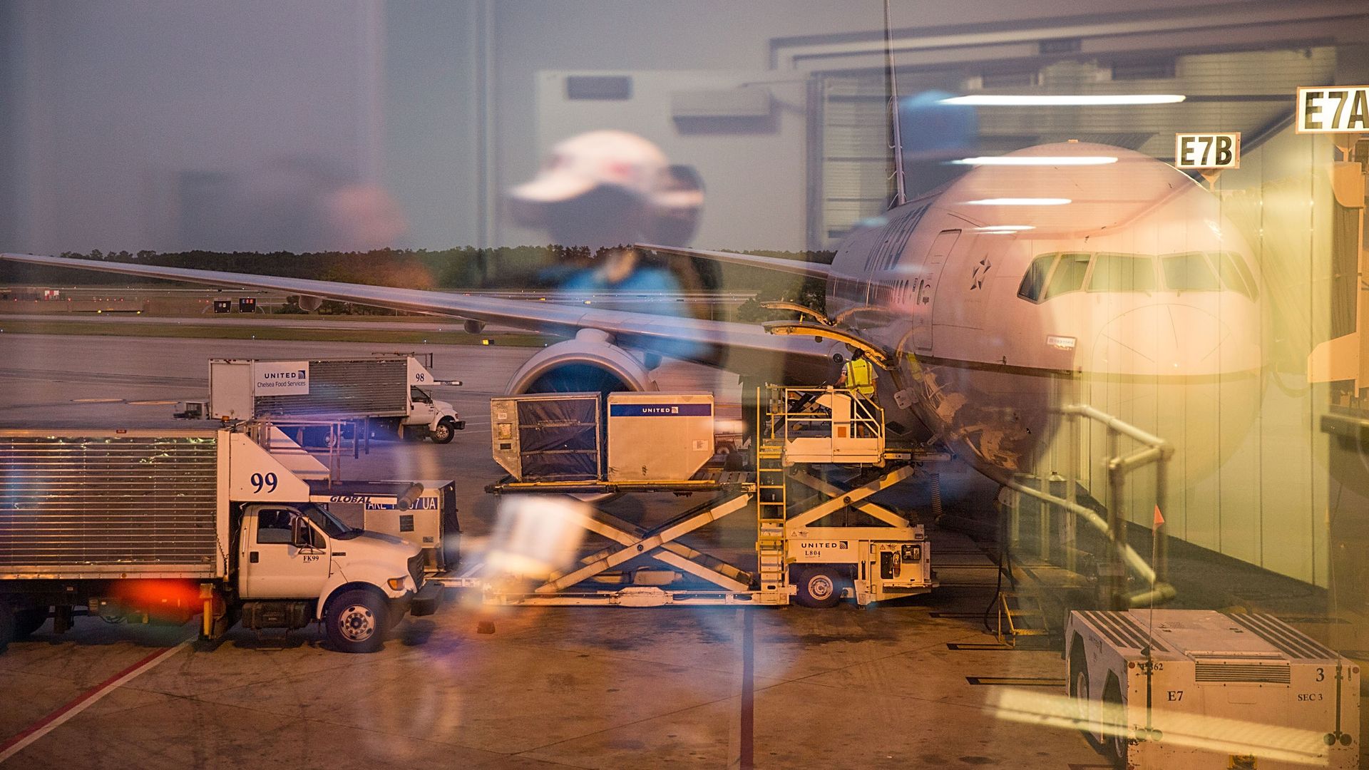 A photo of an airplane parked at a gate taken through a window, with passengers' reflections in the glass