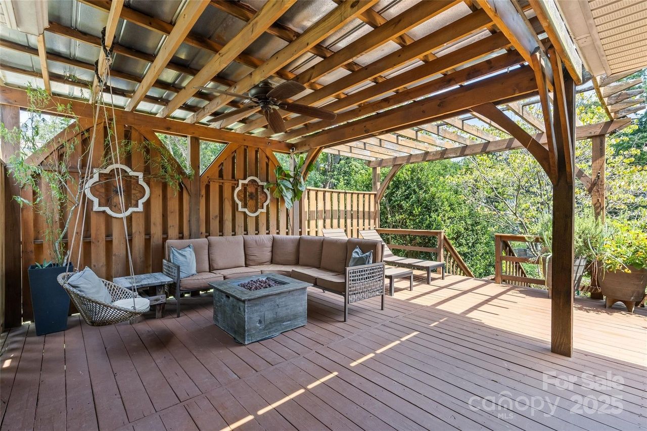 Outdoor wooden deck with sectional sofa, fire pit table, swinging chair, two lounge chairs under a wooden pergola surrounded by greenery.