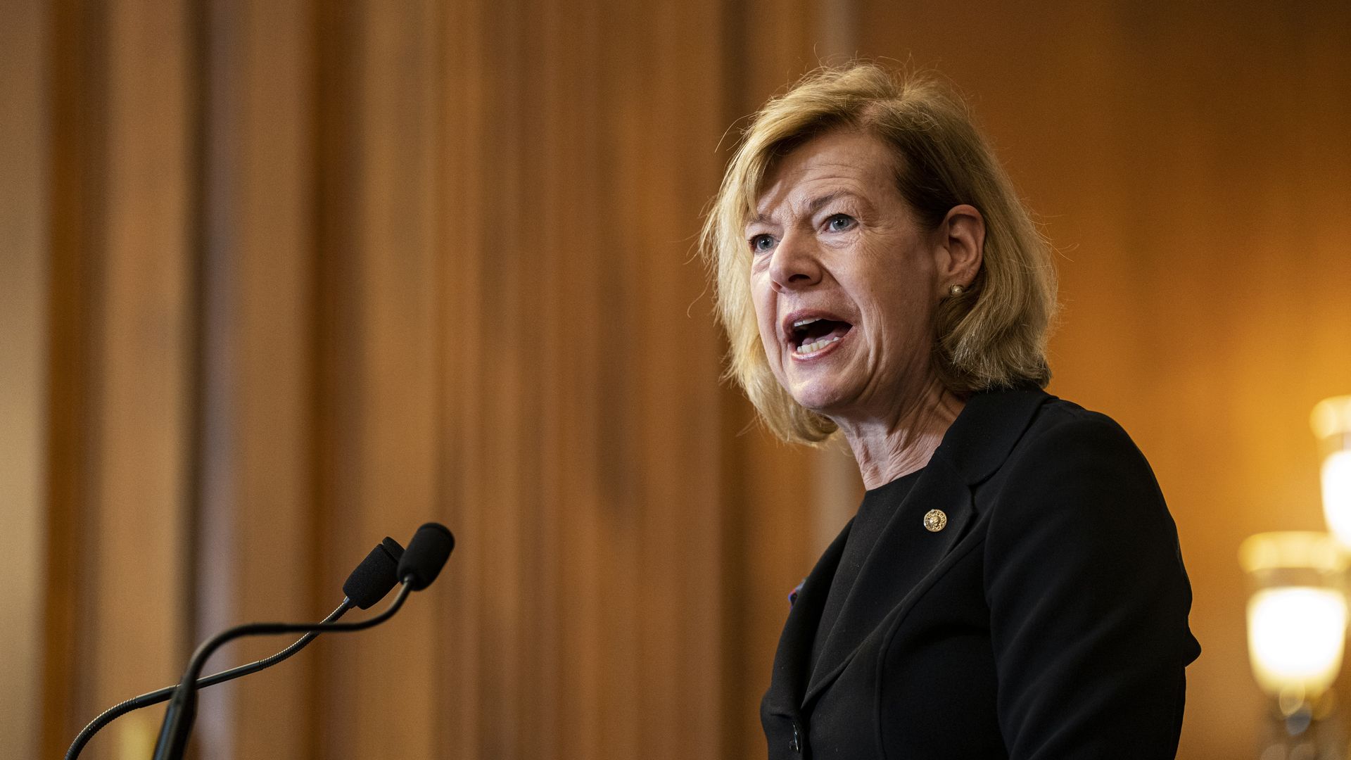 Sen. Tammy Baldwin, wearing a black suit over a black shirt, speaks at a microphone in the Rayburn Room of the Capitol.