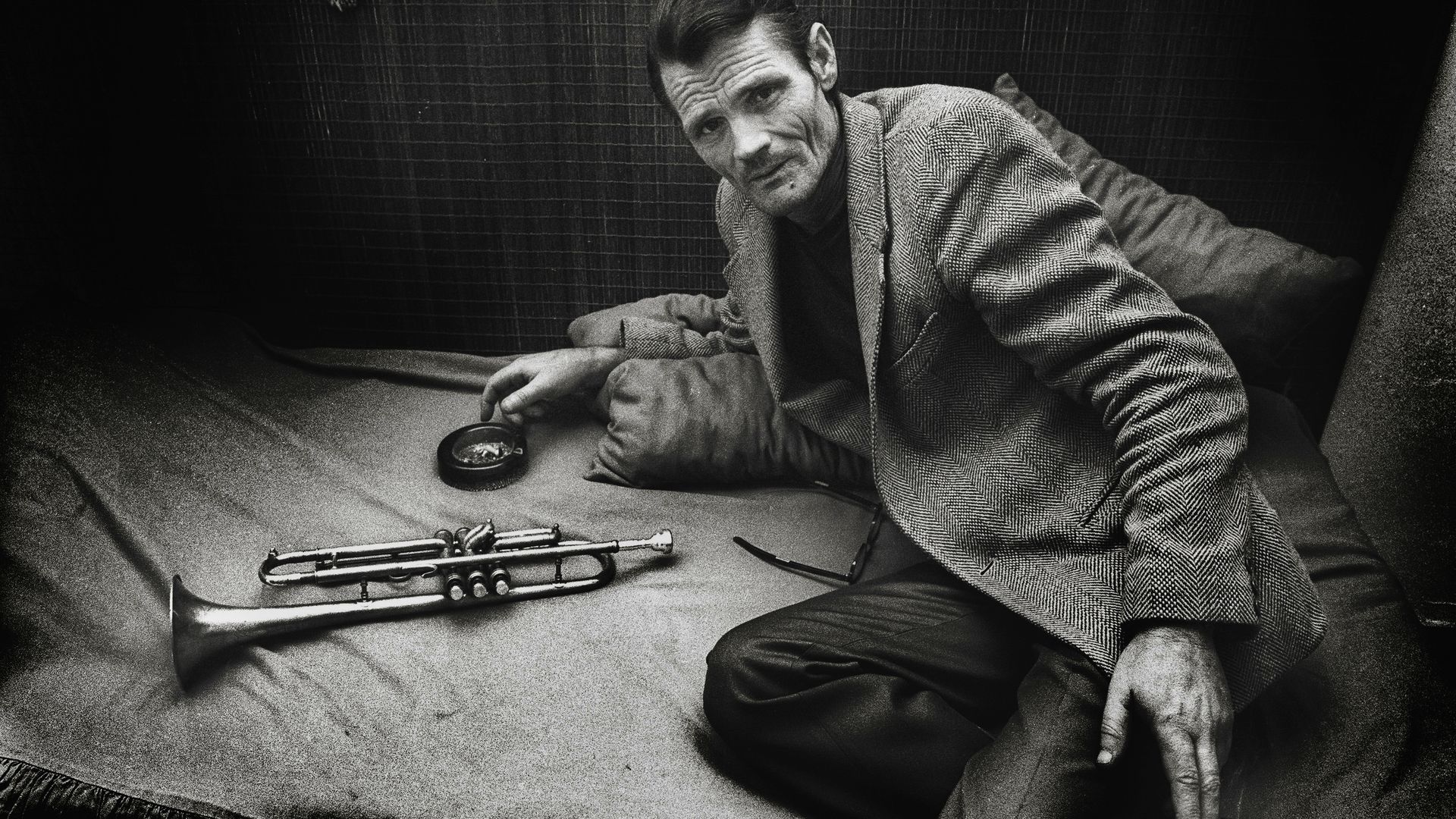 Black-and-white image of a man in a plaid blazer lying on a bed, facing the camera. A trumpet rests on the bed in front, and he holds a cigarette; dark tiled wall behind.