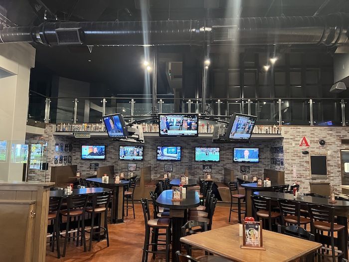 Interior of a sports bar with brick walls, many wall-mounted TVs showing games, a dark ceiling with exposed ducts, and rows of high-top tables and wooden chairs beneath a glass balcony railing.
