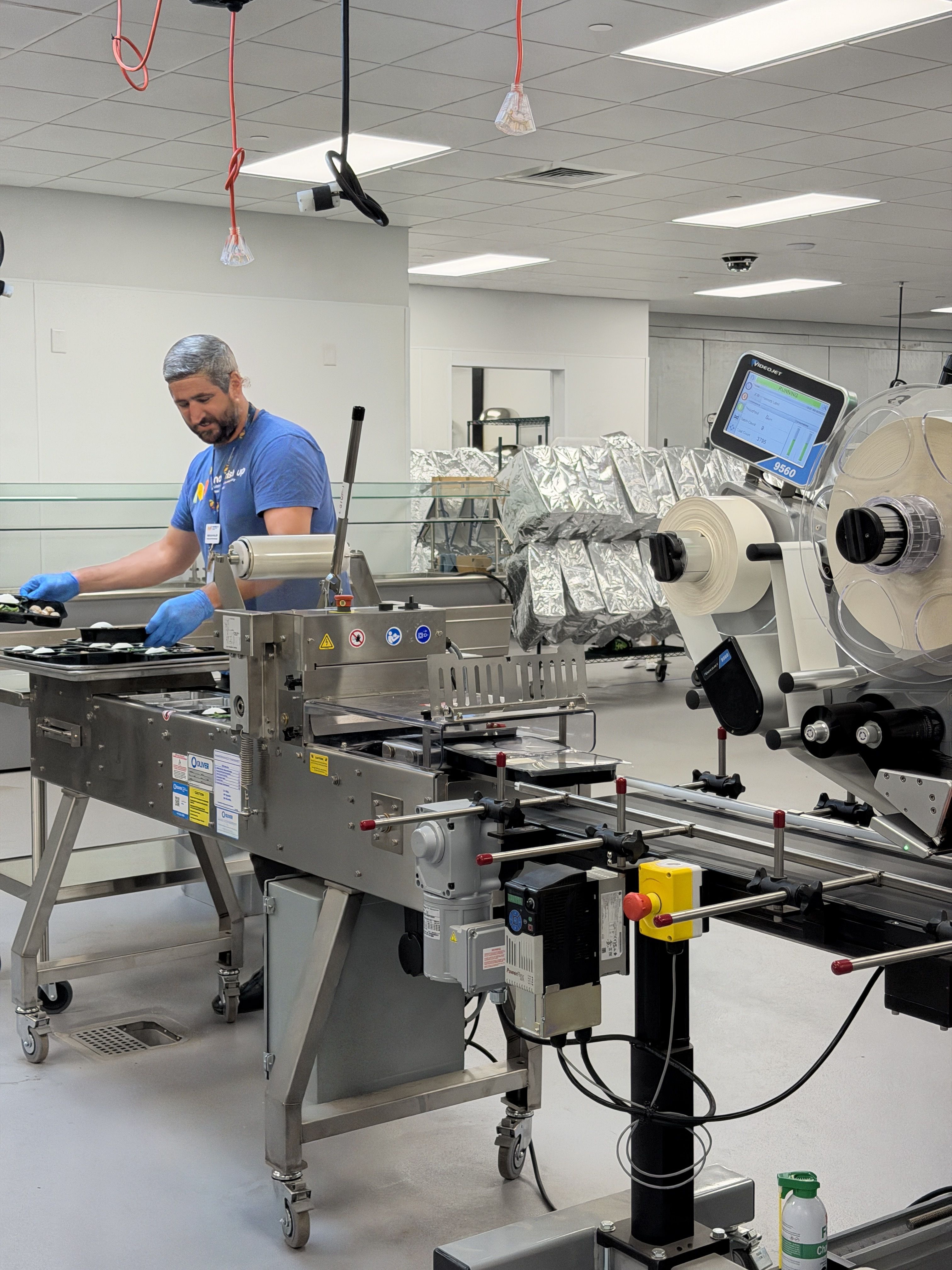 Technician in a blue shirt and gloves operates a stainless steel packaging line in a bright lab. A monitor sits on a large roller machine, with foil-wrapped equipment in the background.