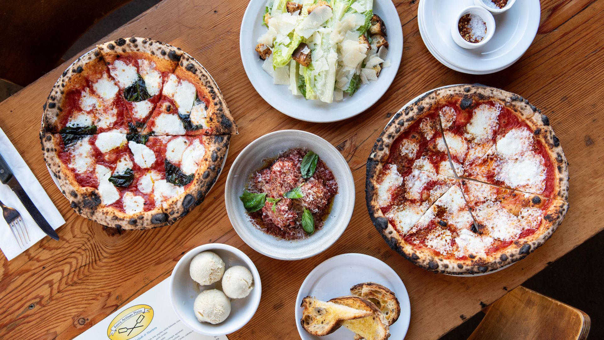 Table with two pizzas with melted cheese and tomato sauce, Caesar salad with shaved Parmesan, bowl of meatballs garnished with basil, plate of toasted bread slices, and two small bowls of chili flakes and salt.