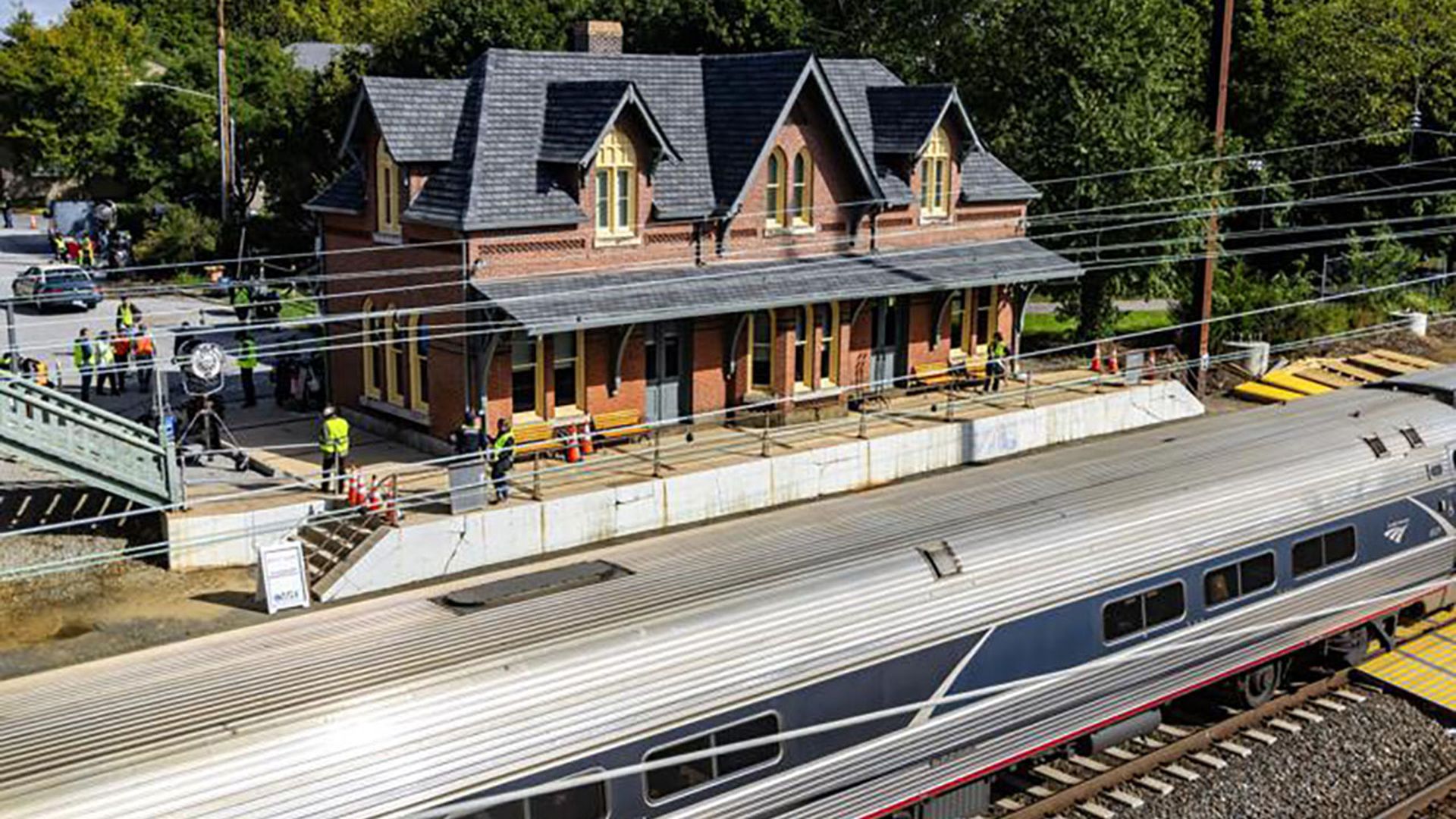Brick train station with a dark slate roof and yellow-trimmed windows sits beside a platform as a silver passenger train runs along tracks; workers in safety vests stand nearby under power lines.
