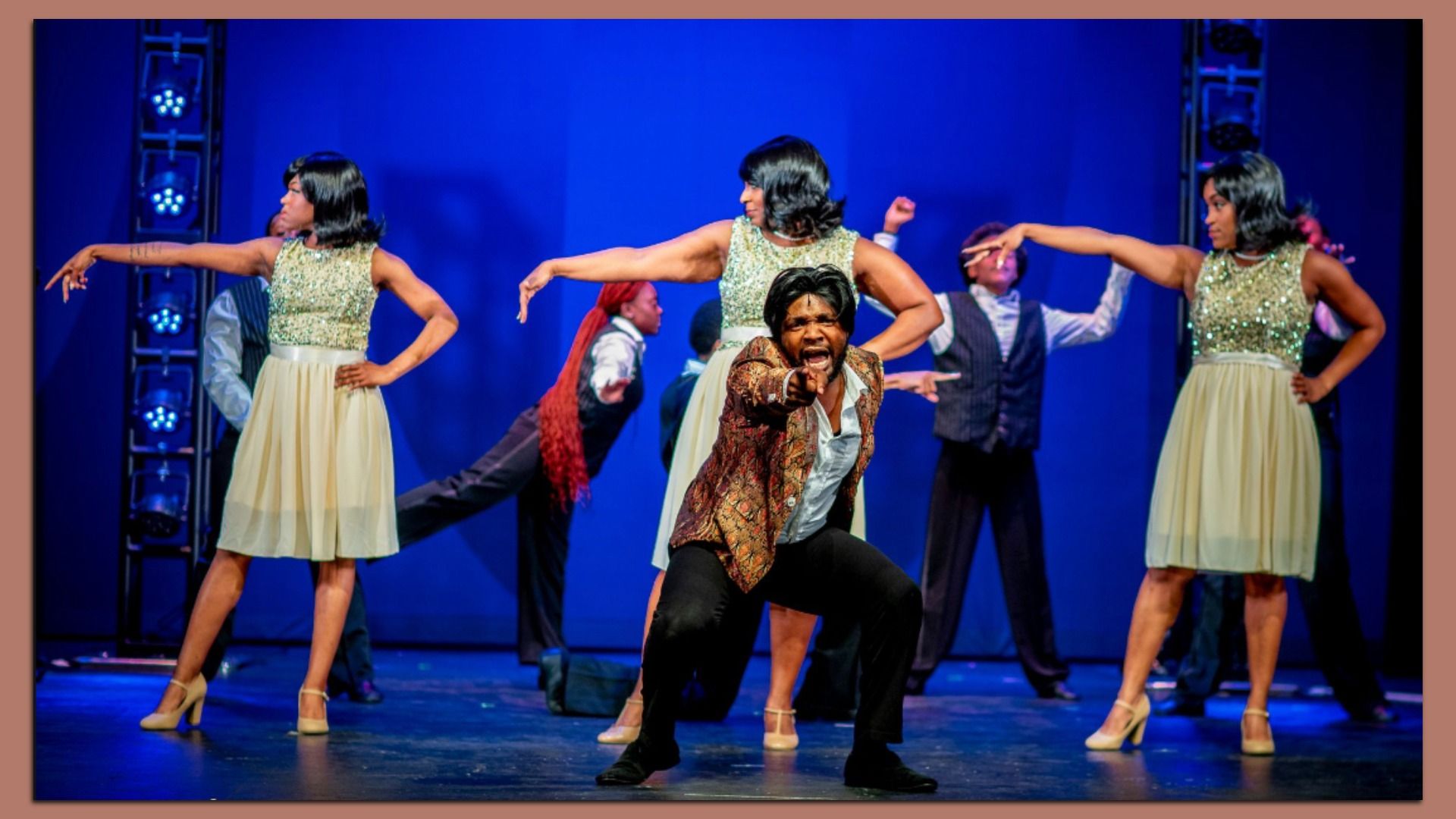 On a blue-lit stage, a man in a patterned brown jacket points at the camera, surrounded by dancers in pale yellow dresses with glittery tops; other performers stand in the background.