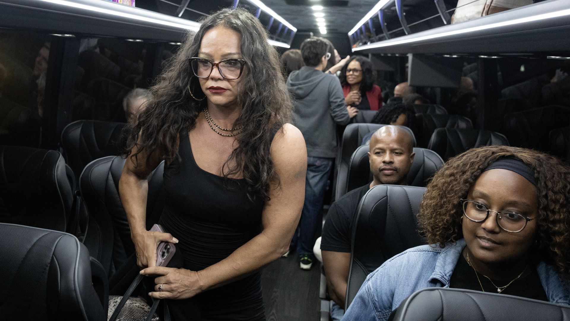 Inside a dimly lit bus, a woman with long curly hair and glasses stands holding a phone and bag. Other passengers sit or stand in black leather seats, some conversing.