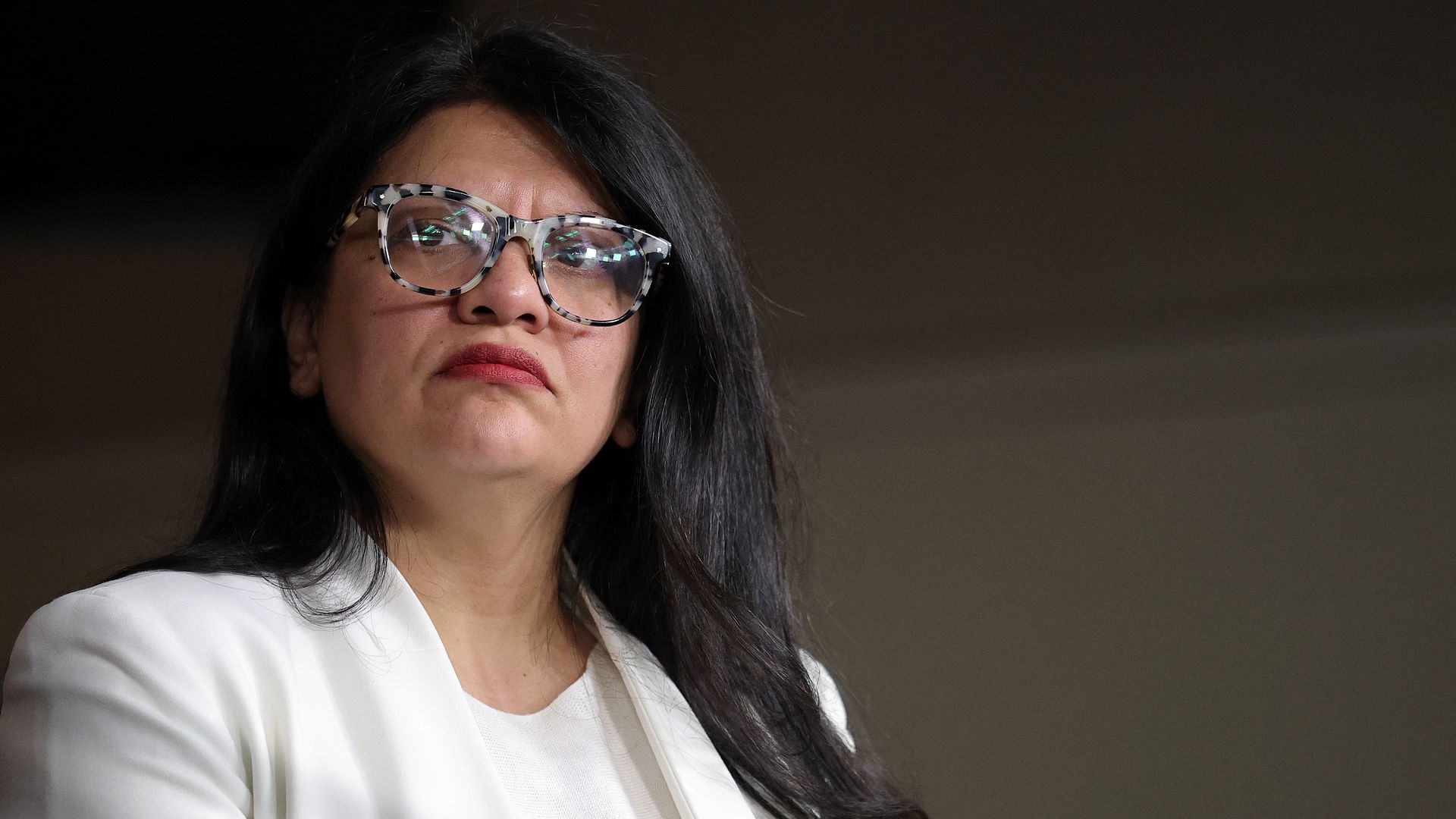 Portrait of a woman with long black hair, wearing patterned rectangular glasses and a white blazer, looking slightly to the left with a neutral expression against a dark blurred background.