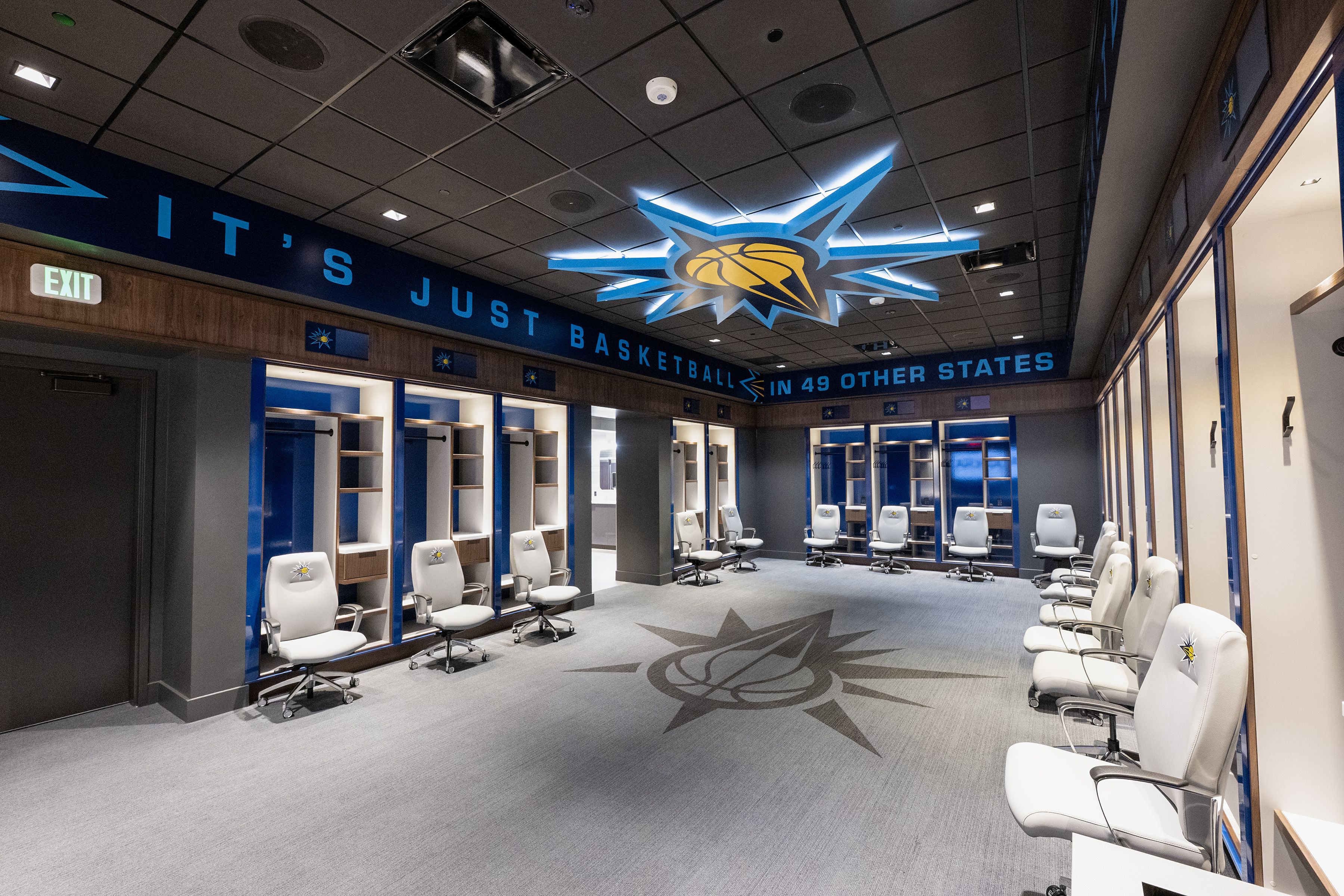 A spacious basketball locker room with white chairs featuring a blue and yellow logo. Blue and wood lockers line the walls, with a large illuminated logo on the ceiling and floor. Text on the wall reads "IT'S JUST BASKETBALL IN 49 OTHER STATES".