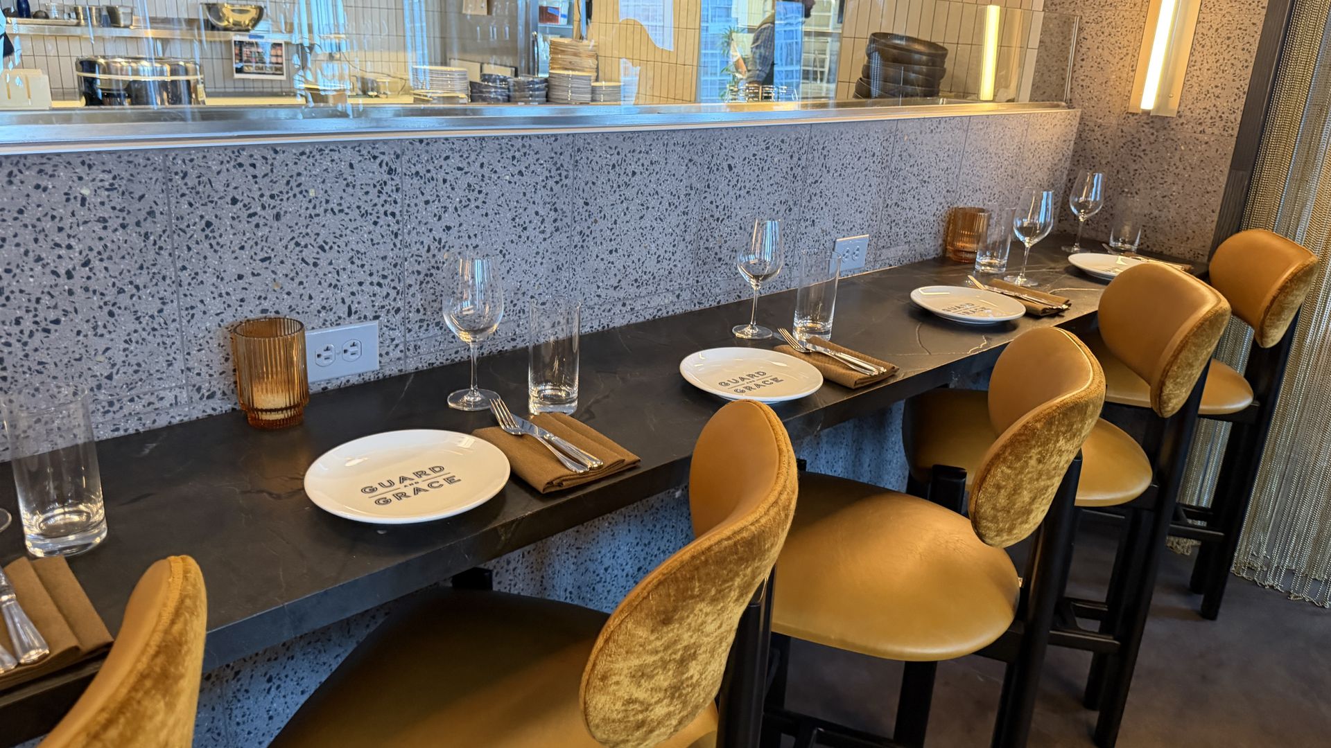 A chic restaurant counter with speckled terrazzo and a glass divider to an open kitchen. Table settings include napkins, plates with text, wine and water glasses, and tan velvet chairs along bar.