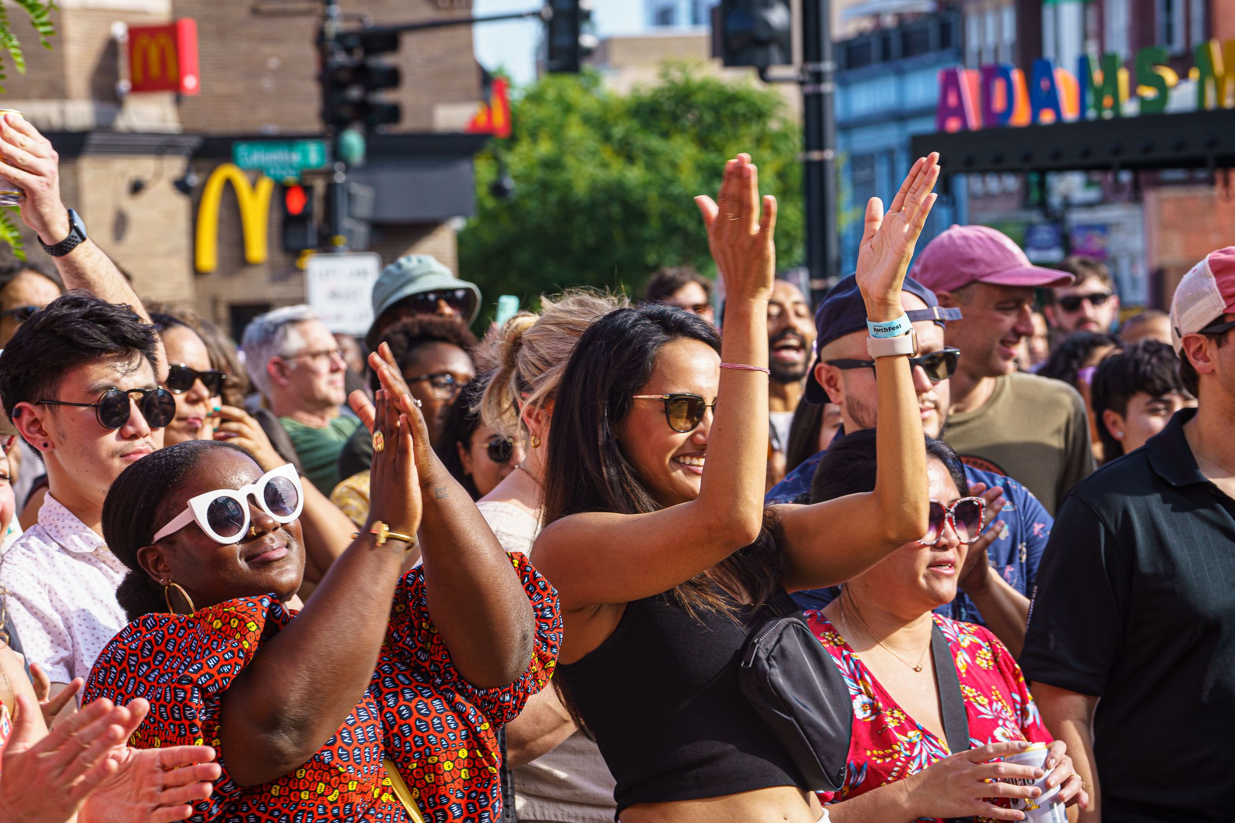 Diverse crowd of people outdoors clapping and enjoying an event on a sunny day, with McDonald's and Adams sign visible in the background.