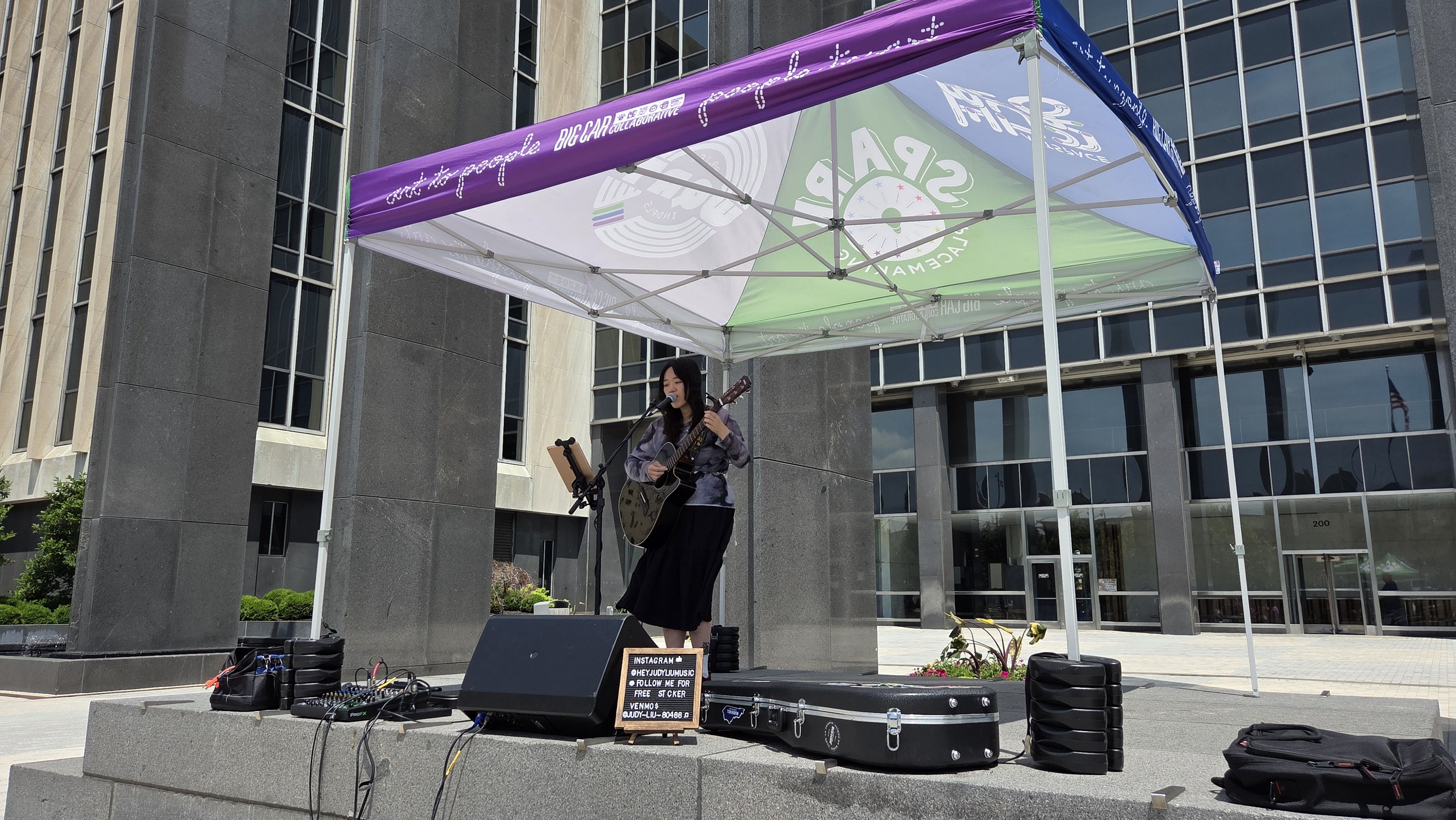 A female guitarist sings into a microphone on an outdoor stage under a purple canopy, with guitar, music stand, and gear on the platform in front of a modern glass building.