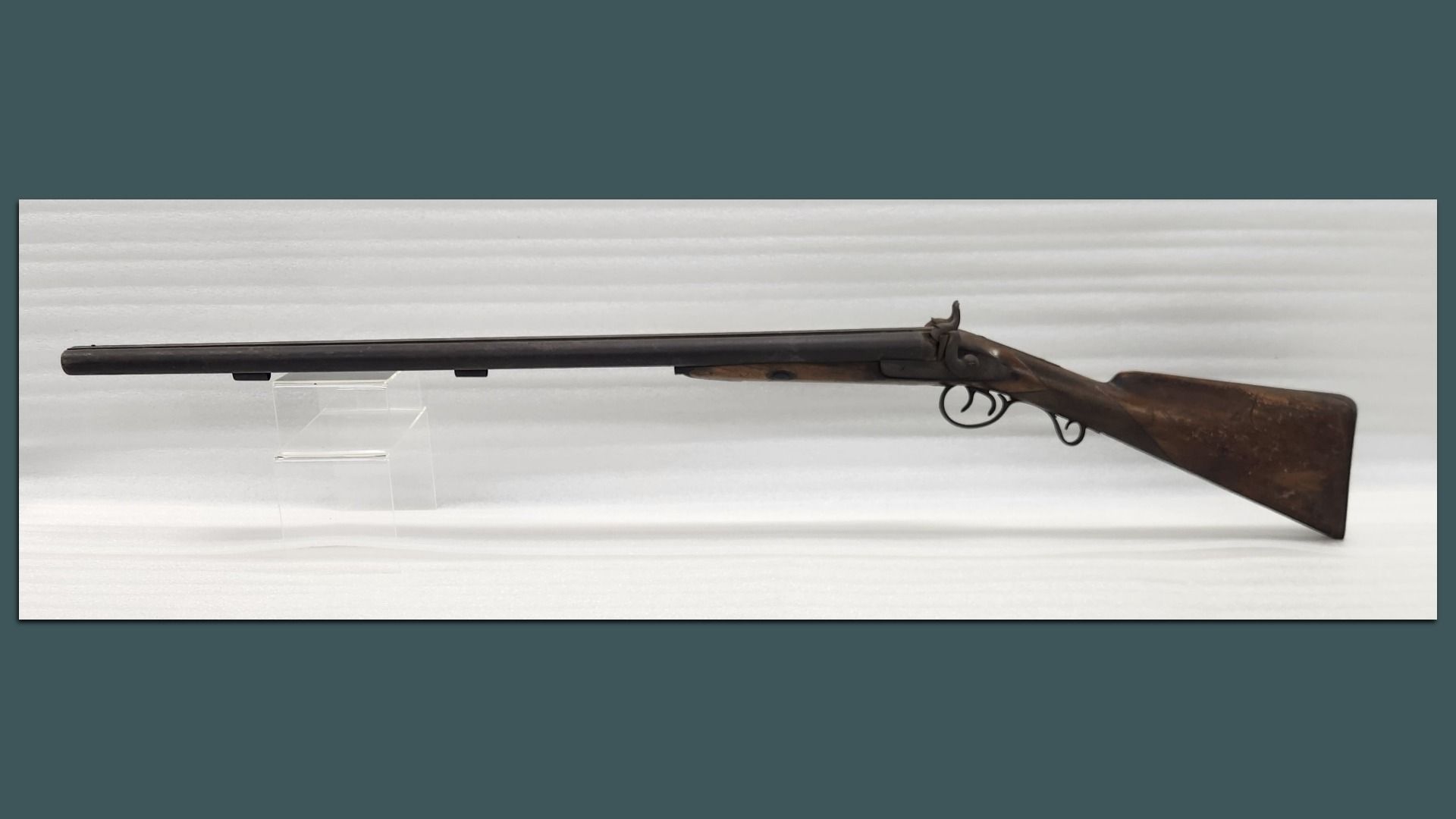 Side view of an antique muzzle-loading rifle with a long dark barrel, wooden stock, and curved trigger guard, displayed on a clear stand against a light gray backdrop.