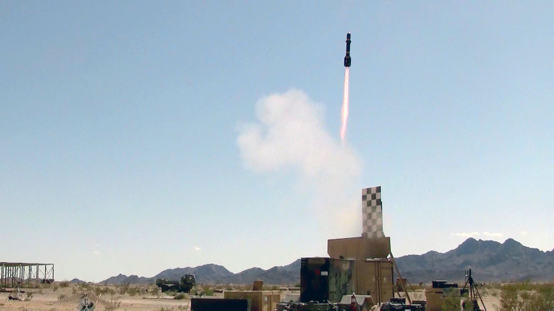 A missile launches vertically from a desert launch site, with a pink-orange flame trailing into a clear blue sky.