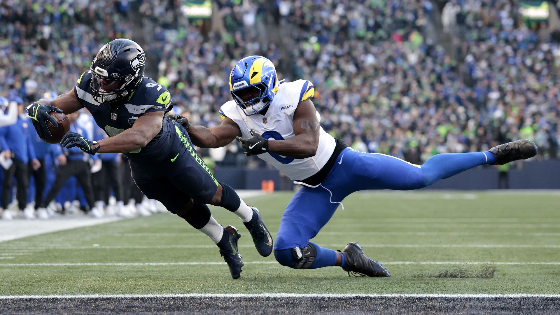 A Seattle Seahawks player in a dark blue jersey with green lettering launches into the endzone holding a football while a Rams player in a white jersey and light blue pants attempts to stop the Seahawks player from behind. A full stadium is visible in the background, blurred. 