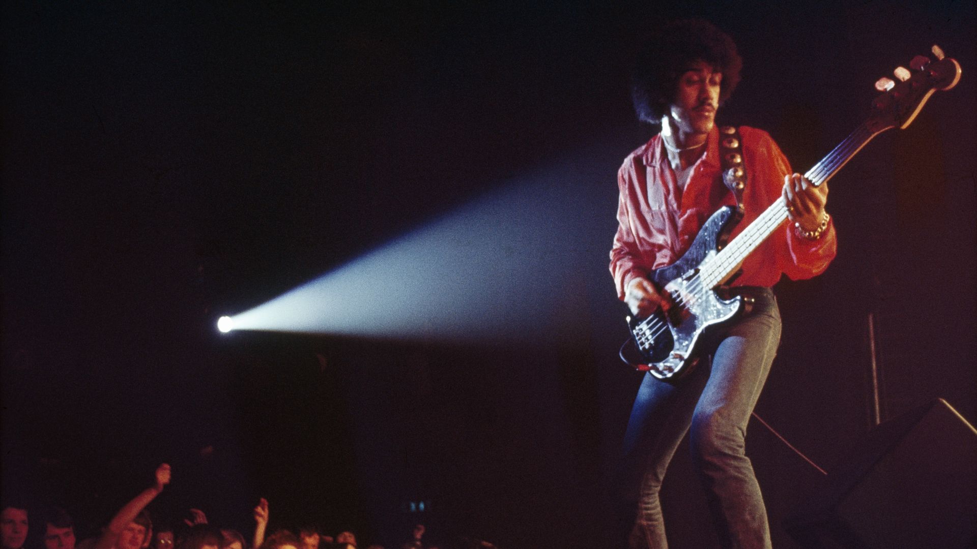 An afro-haired musician in a red shirt plays a silver bass on a dark stage, bathed in a white spotlight from the left, while a cheering crowd fills the foreground.