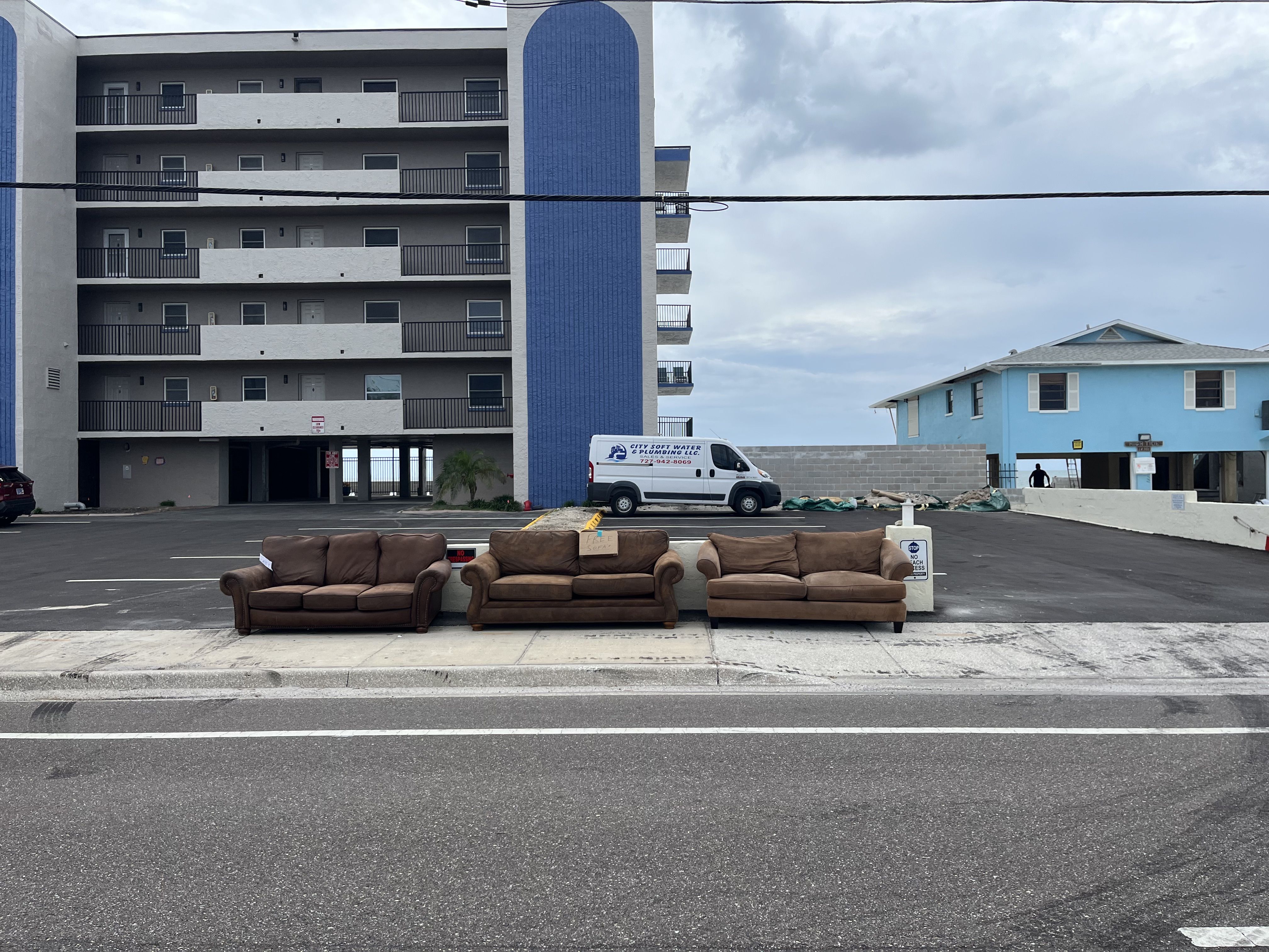 Three brown sofas placed on a sidewalk in front of a parking lot, with a tall white and blue apartment building and a blue house in the background under a cloudy sky.