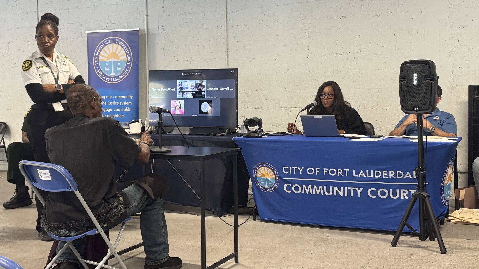 Security officer in uniform stands left as Judge Florence Barner at a table with a Fort Lauderdale Community Court banner; a monitor shows a video call, a microphone, a laptop, and a large speaker nearby. A man sits in front of the judge.