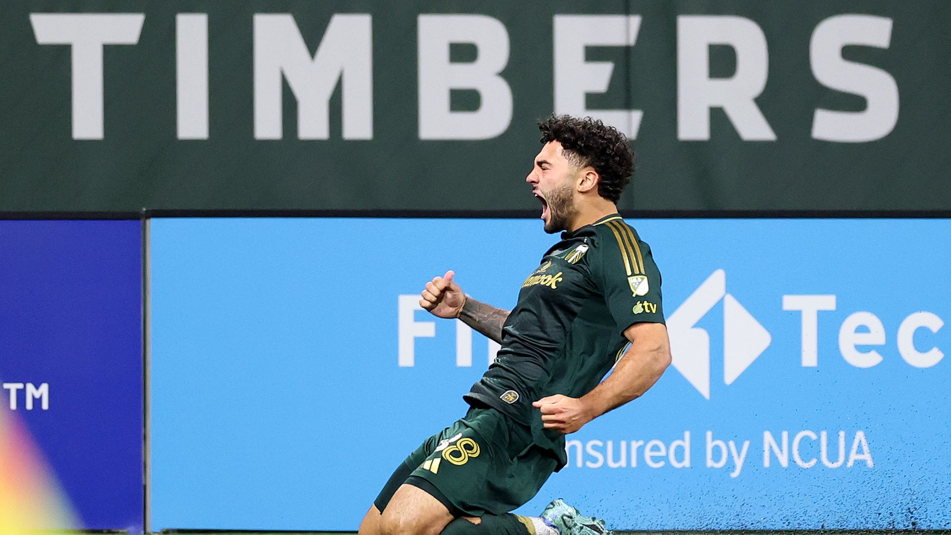 Soccer player in green Portland Timbers uniform celebrates by sliding on knees on field, fists clenched, mouth open shouting, with large TIMBERS sign in background.