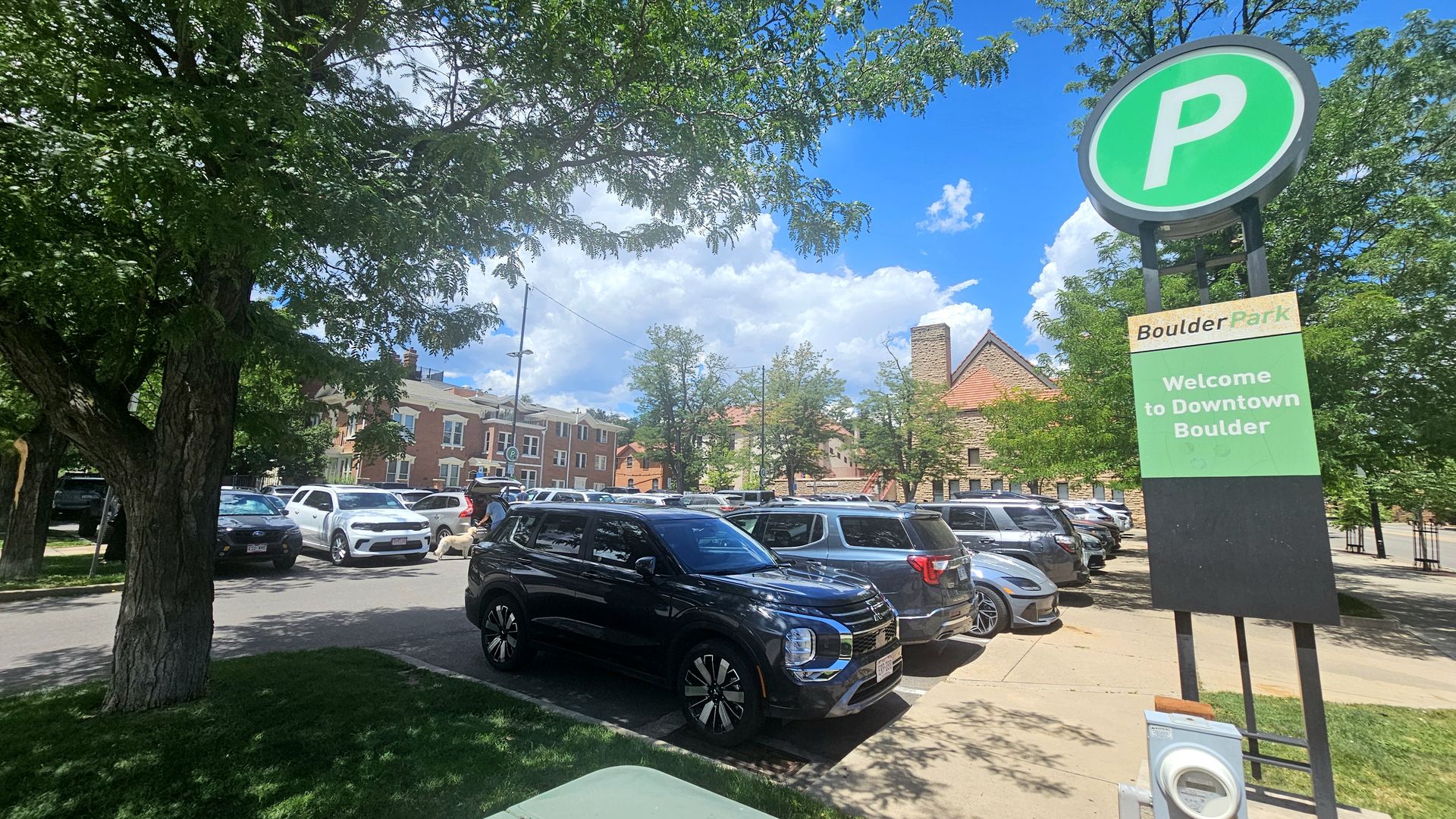 Sunny parking lot framed by trees, with a large green 'P' sign to the right and a sign reading "Boulder Park—Welcome to Downtown Boulder". Cars line the lot under a bright blue sky.