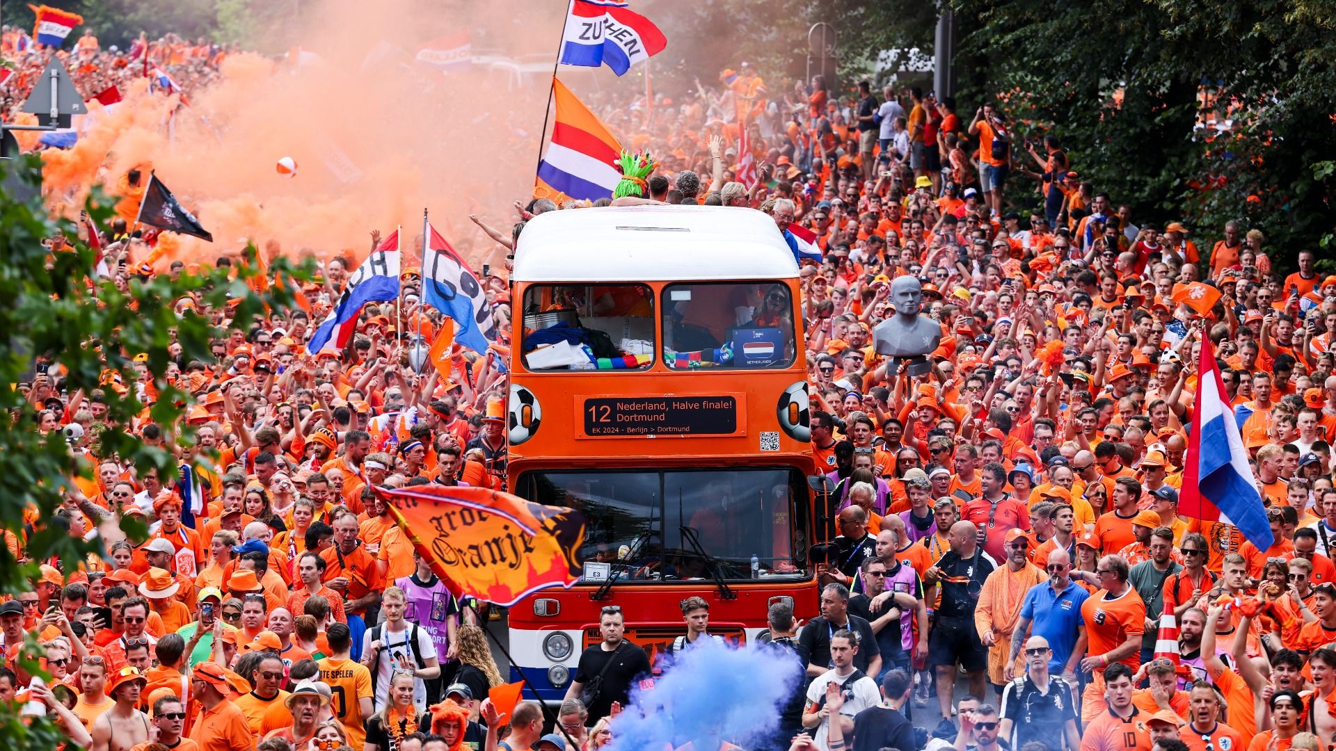 Dutch fans surrounding an orange bus marching down the street with flags and colored smoke, celebrating before a match against England in 2024 in Dortmund. 