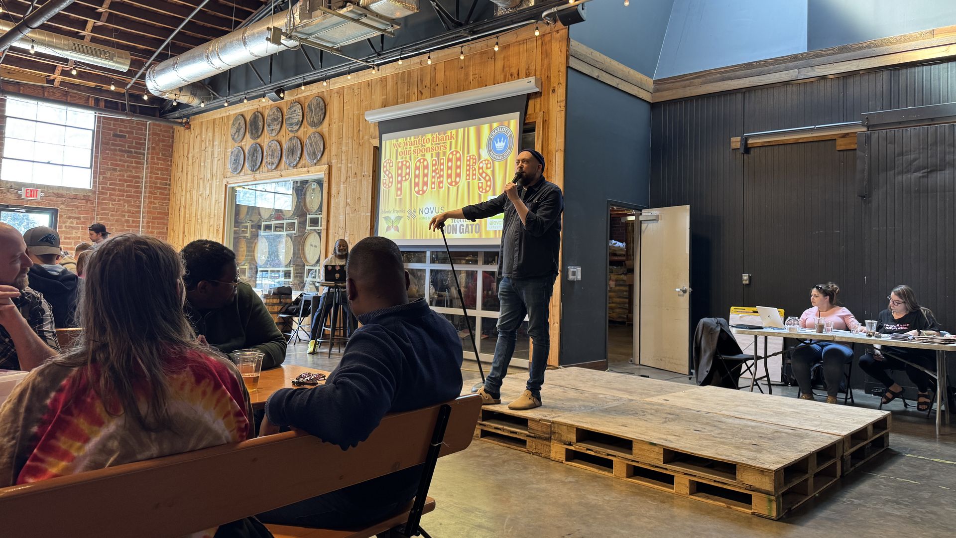 Man with microphone stands on wooden pallet stage in a rustic room with wood walls and barrel decor, speaking to seated audience at a casual indoor event with a sponsor slide projected.