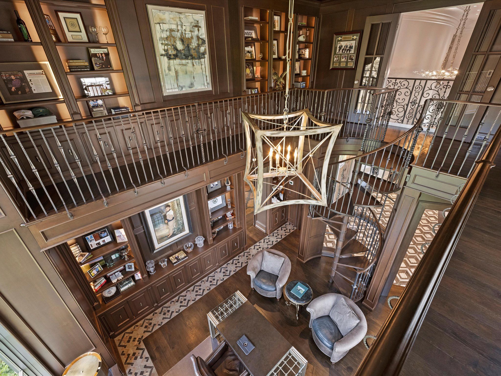 Luxurious two-story home library with dark wood paneling, built-in shelves, spiral staircase, modern chandelier, two gray armchairs, and a large desk with leather chair.