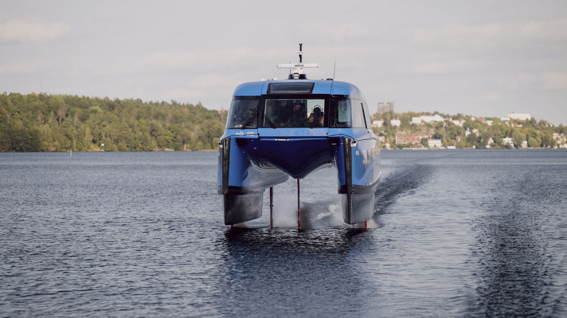 A sleek blue passenger boat on calm water, viewed from the front. The vessel has a glass cockpit, a radar atop, and a distant shoreline with trees and buildings in the background.