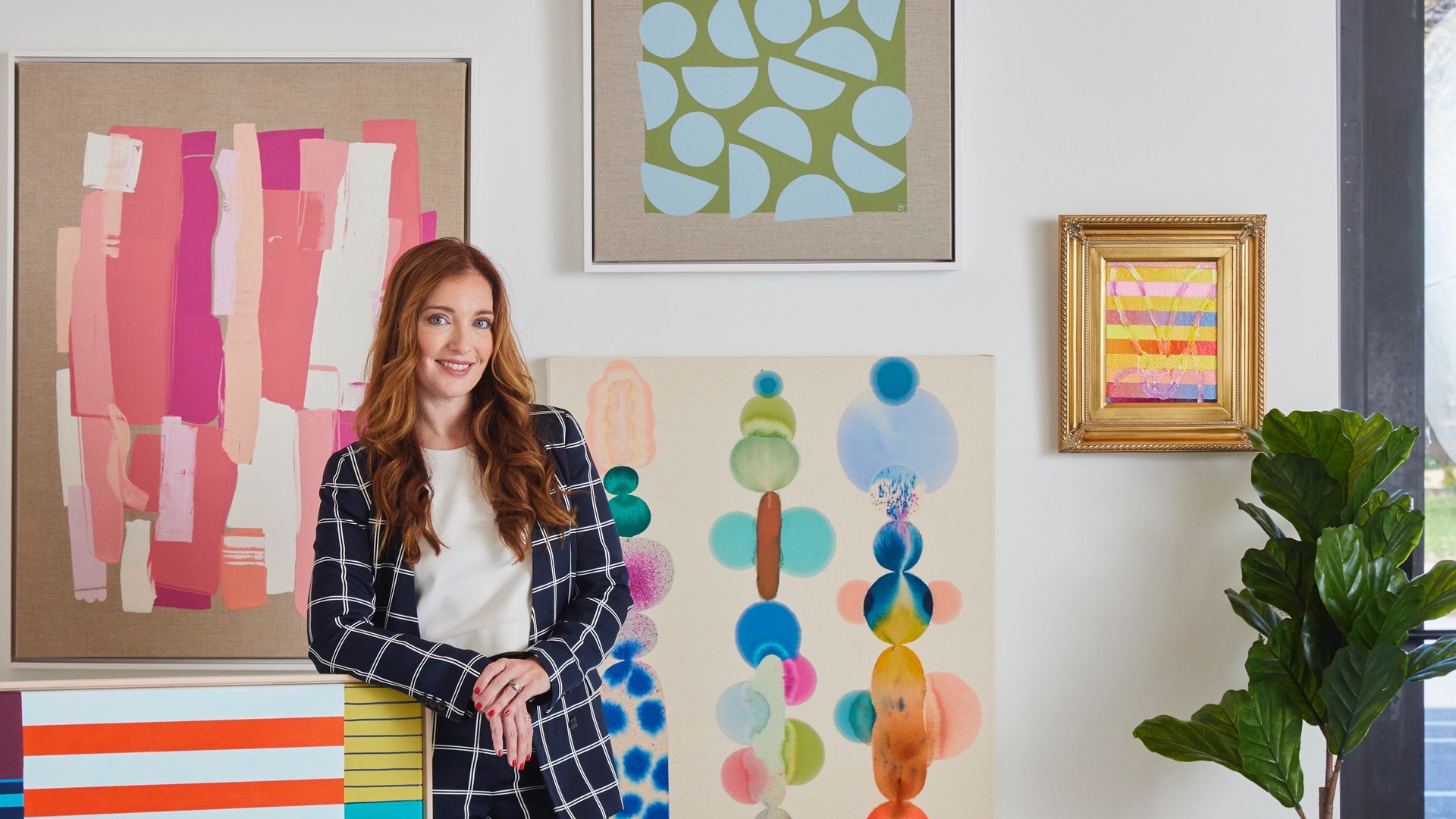 Liz Lidgett in navy plaid suit and burgundy heels leaning on colorful striped artwork in a bright room with abstract paintings, green plants, and large windows.
