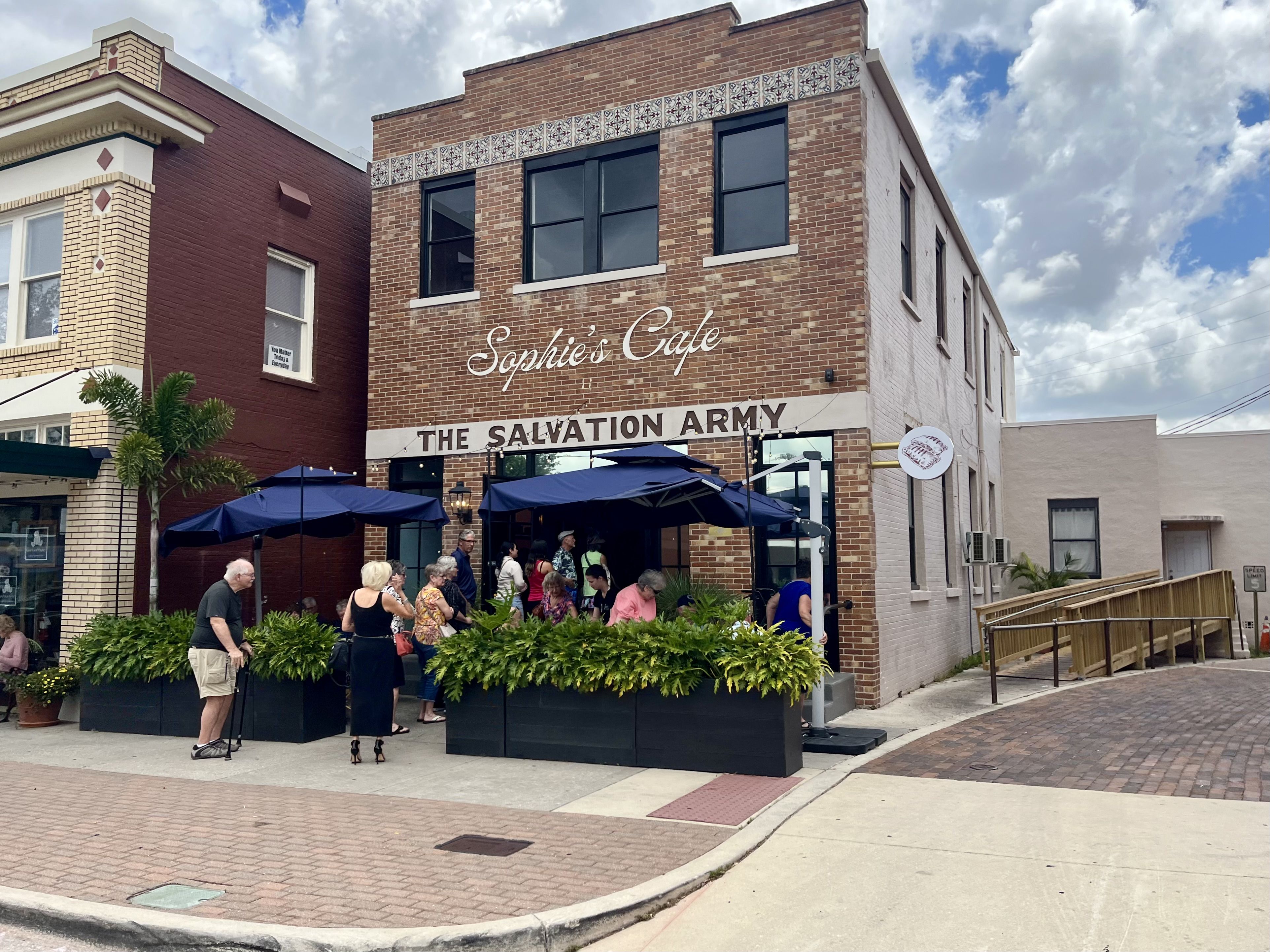 A line forms outside a brick building that says in white cursive "Sophie's Cafe" and, below that, "THE SALVATION ARMY."