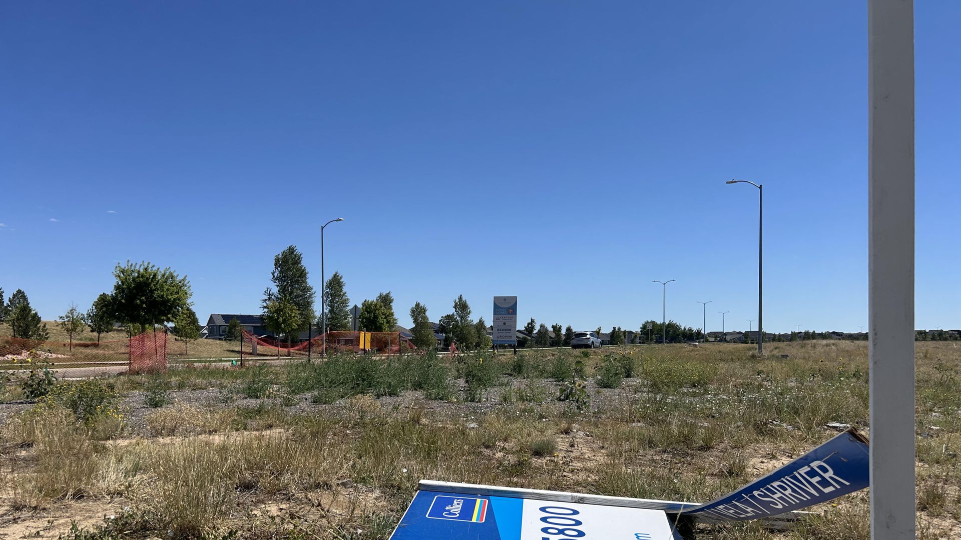 Bright blue sky over a dry, grassy lot with scattered trees, orange construction fencing, and streetlights. A large blue-and-white sign lies overturned in the foreground near a tall white post.