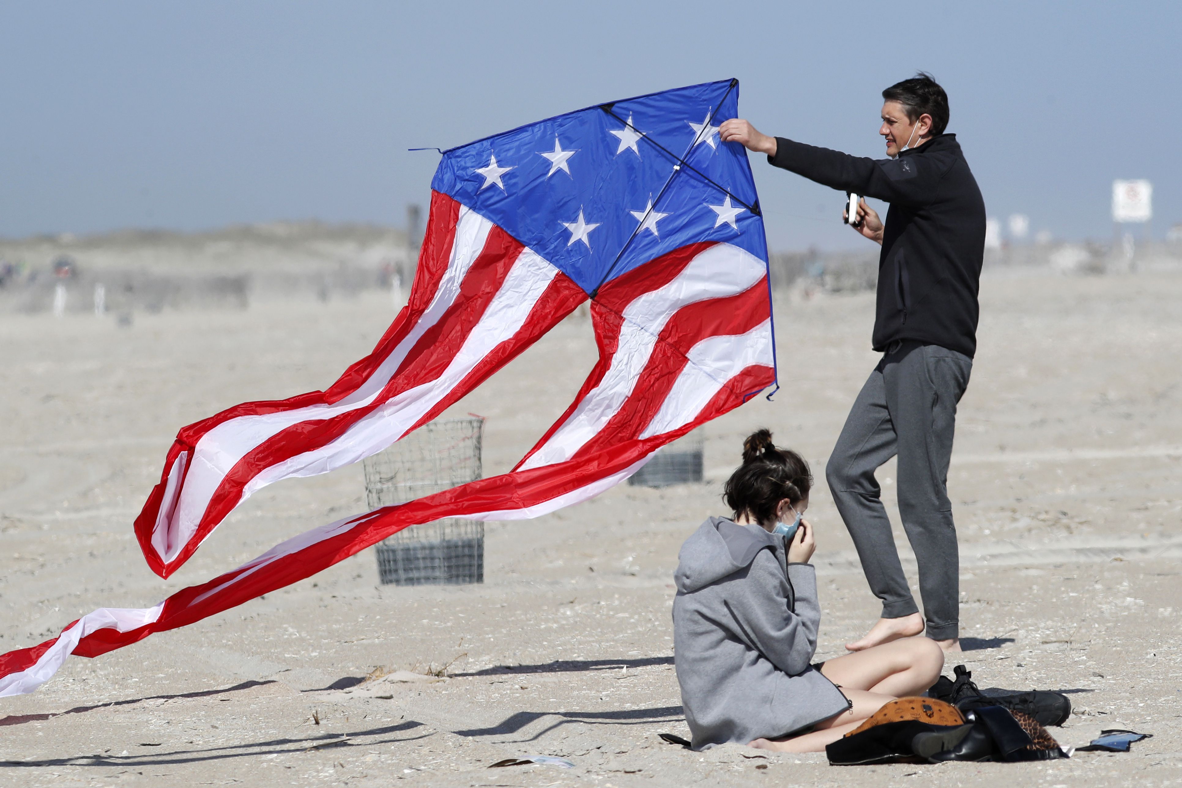 In this image, a man holds a kite on a beach as a woman sits next to him 