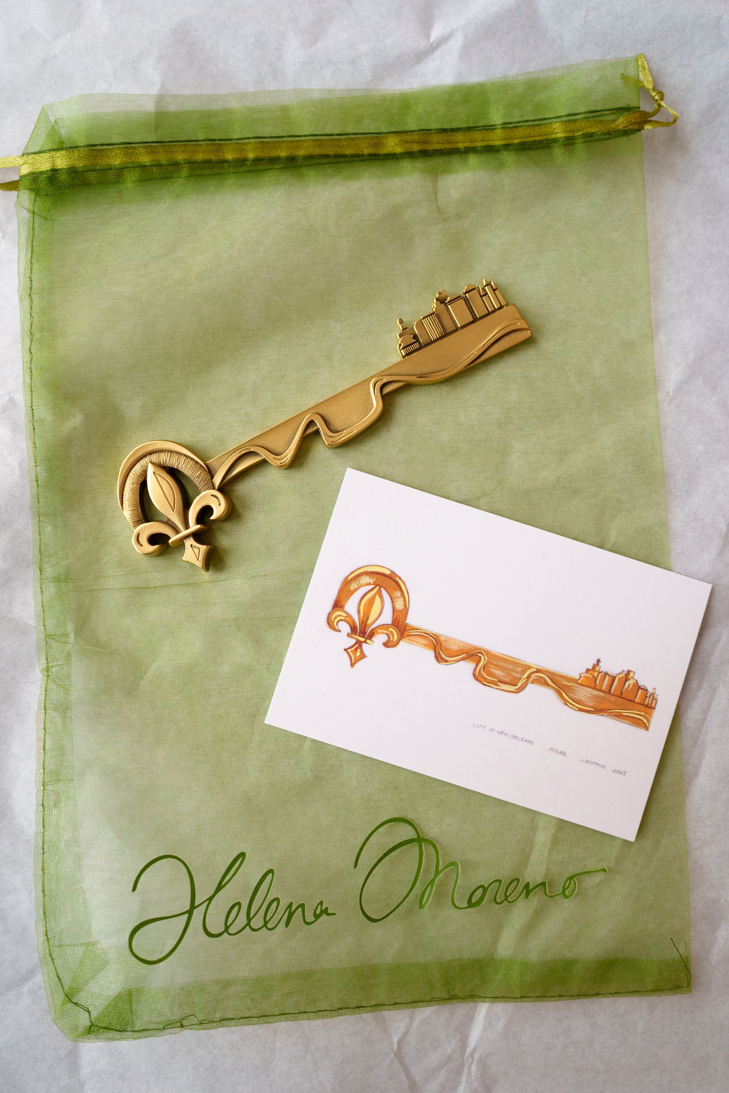Light-green translucent drawstring bag with a gold ribbon, an ornate gold key-shaped ornament, and a white card showing an orange-brown curved-key illustration with a city skyline.