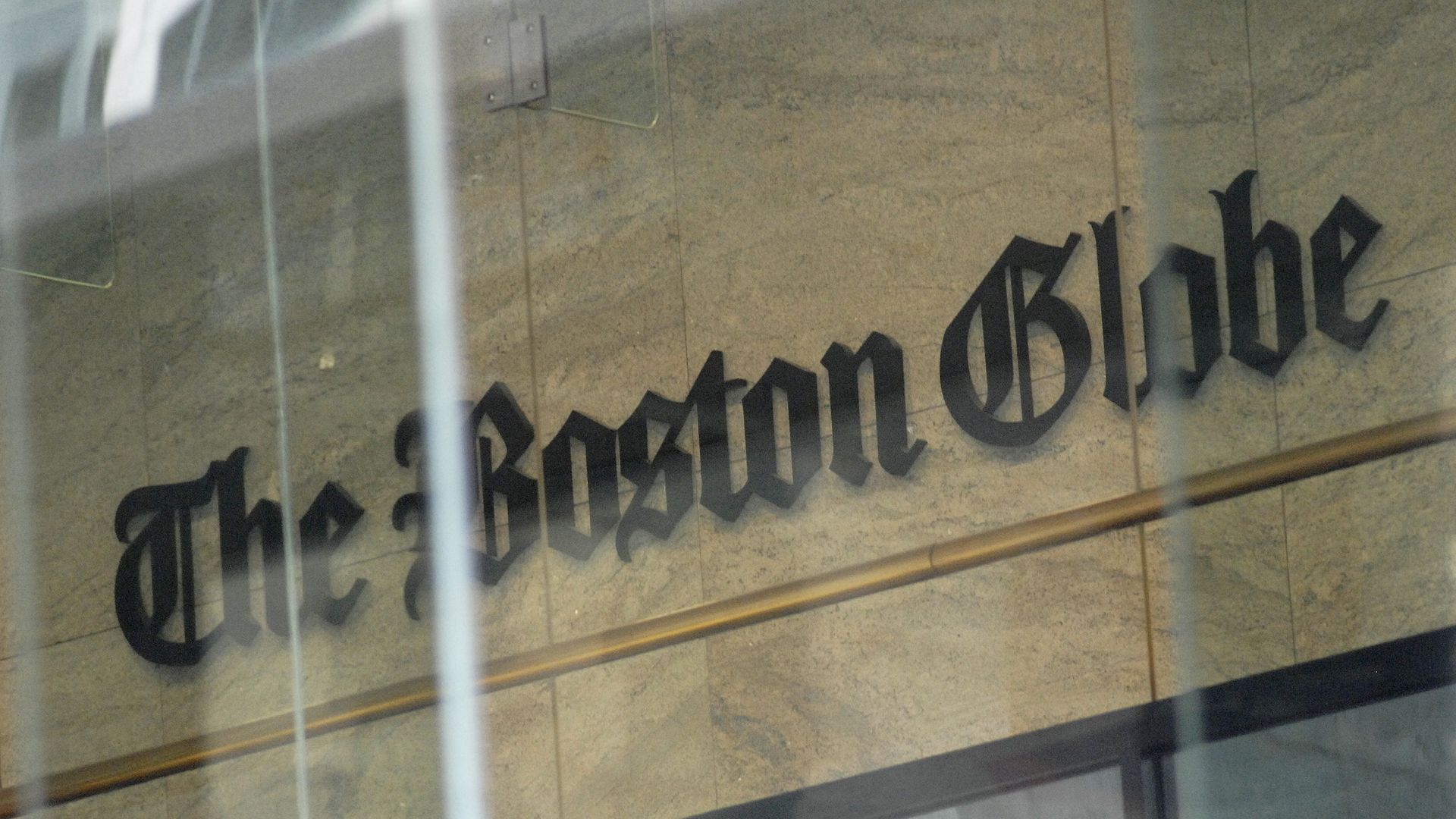 Black Gothic-style lettering of "The Boston Globe" on a beige stone wall, seen through vertical reflective glass panels.