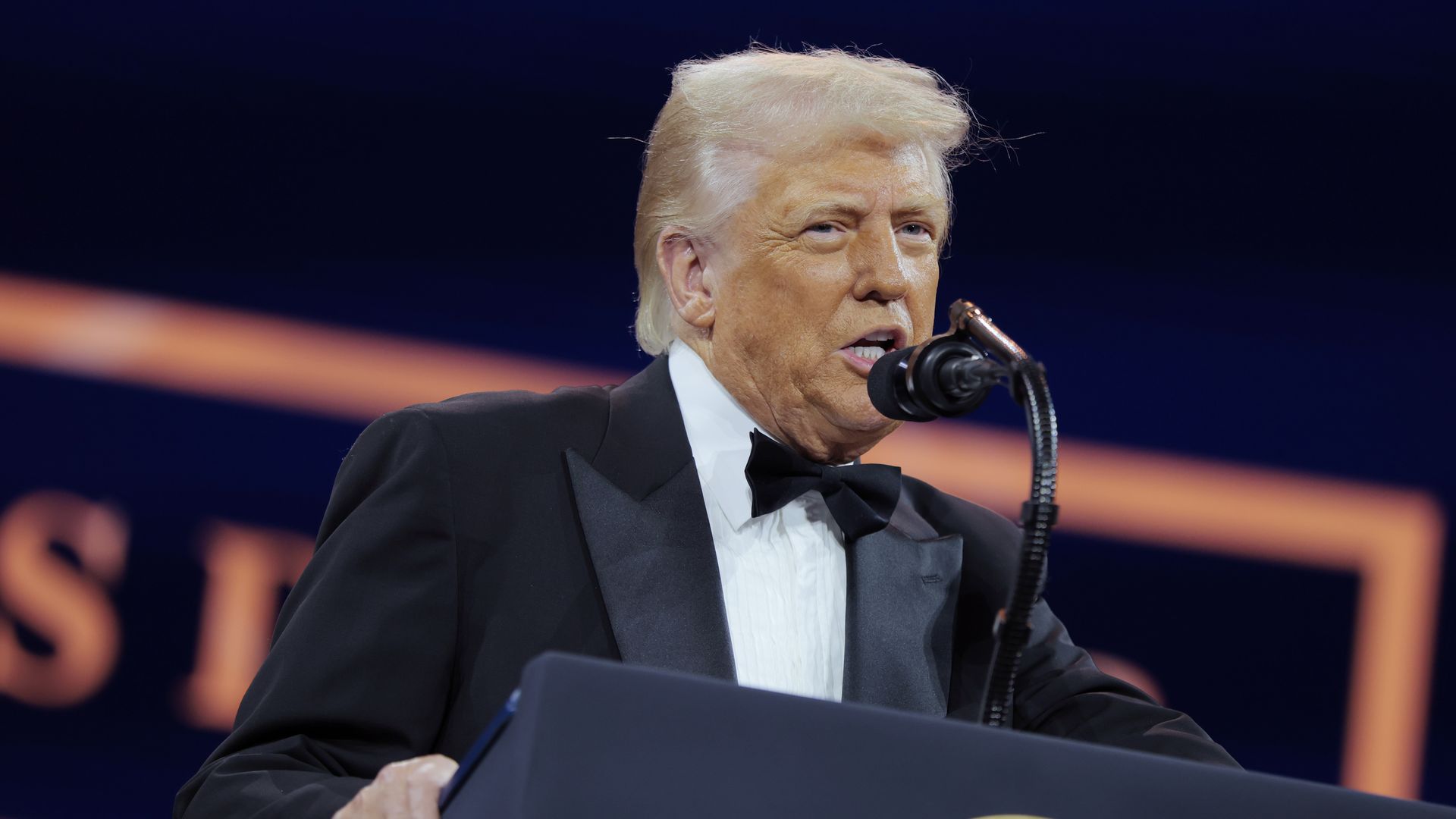 Donald Trump wearing a tuxedo and speaking into microphones at a podium with the presidential seal on it while standing in front of a blue and orange backdrop.