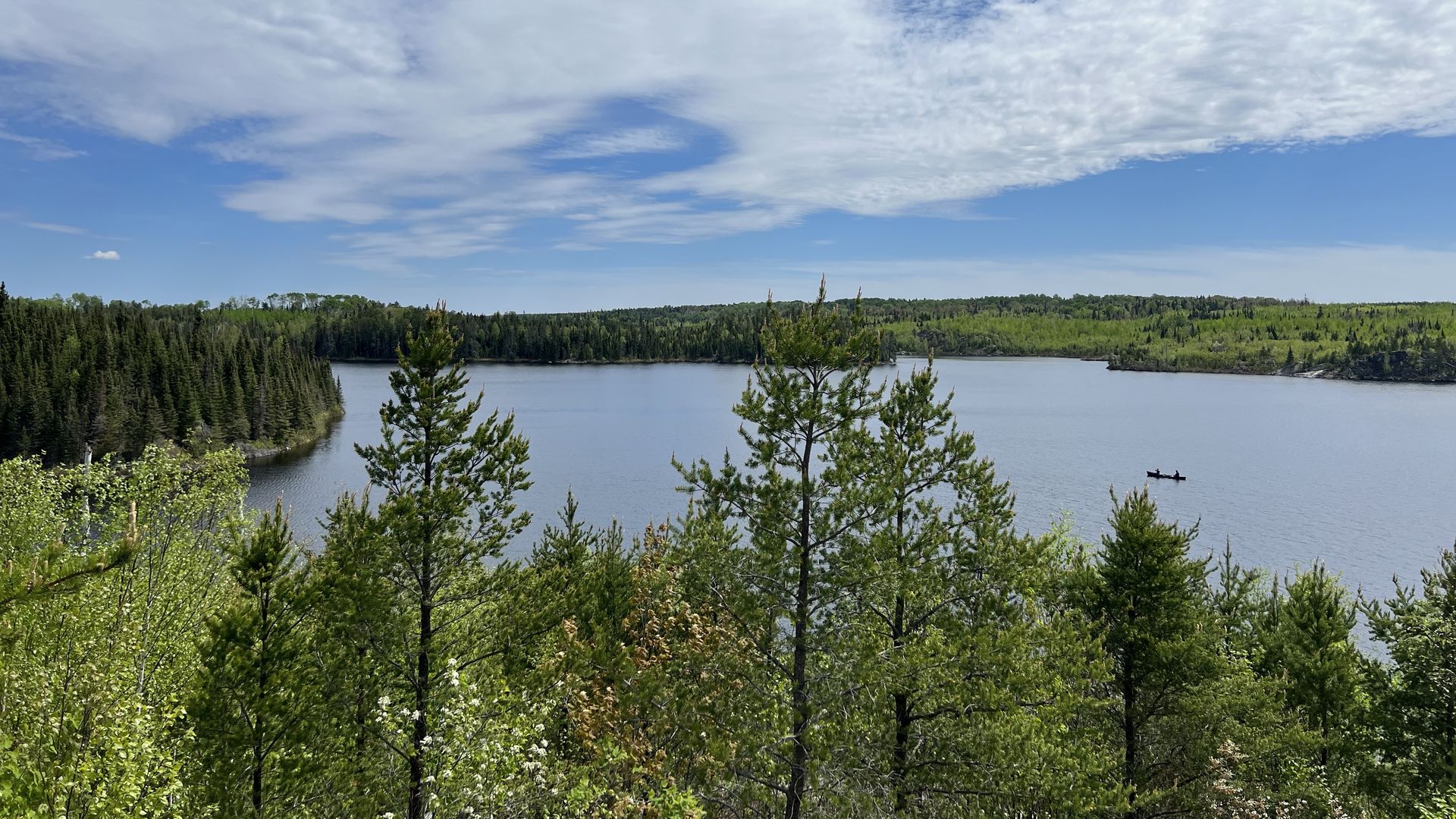 A shot taken from the top of a hill overlooking a tree-ringed lake under blue skies with a small canoe traveling on the water below