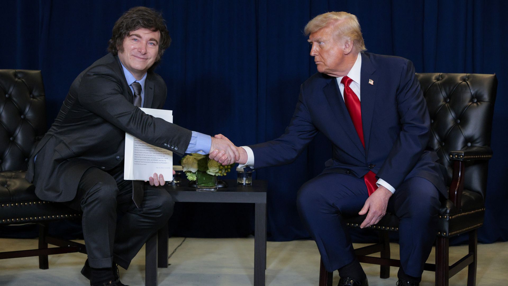 Two men, seated side by side, shake hands, the man on the left holding up documents for a camera. 