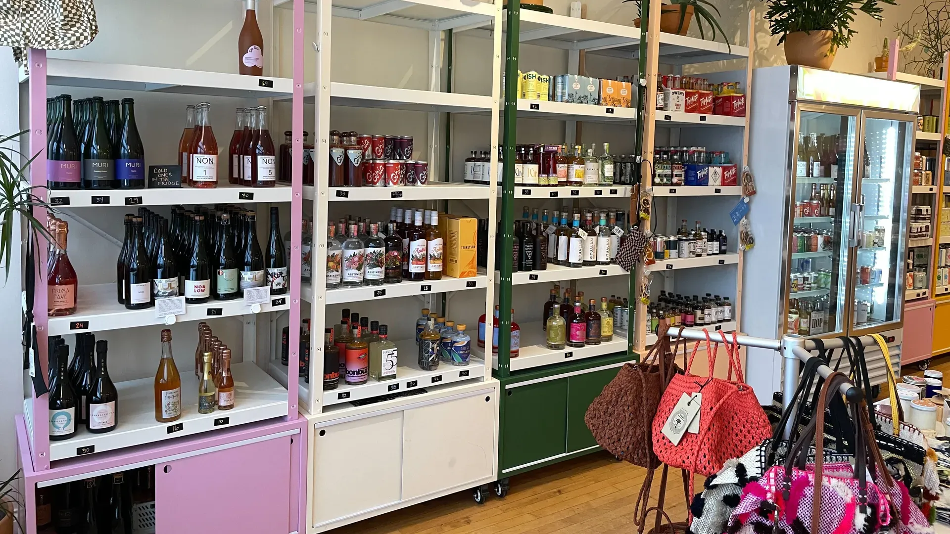 Interior of a colorful shop with white and pastel pink shelves filled with wine bottles, spirits, and soft drinks; a glass-door fridge on the right and bags in the foreground.