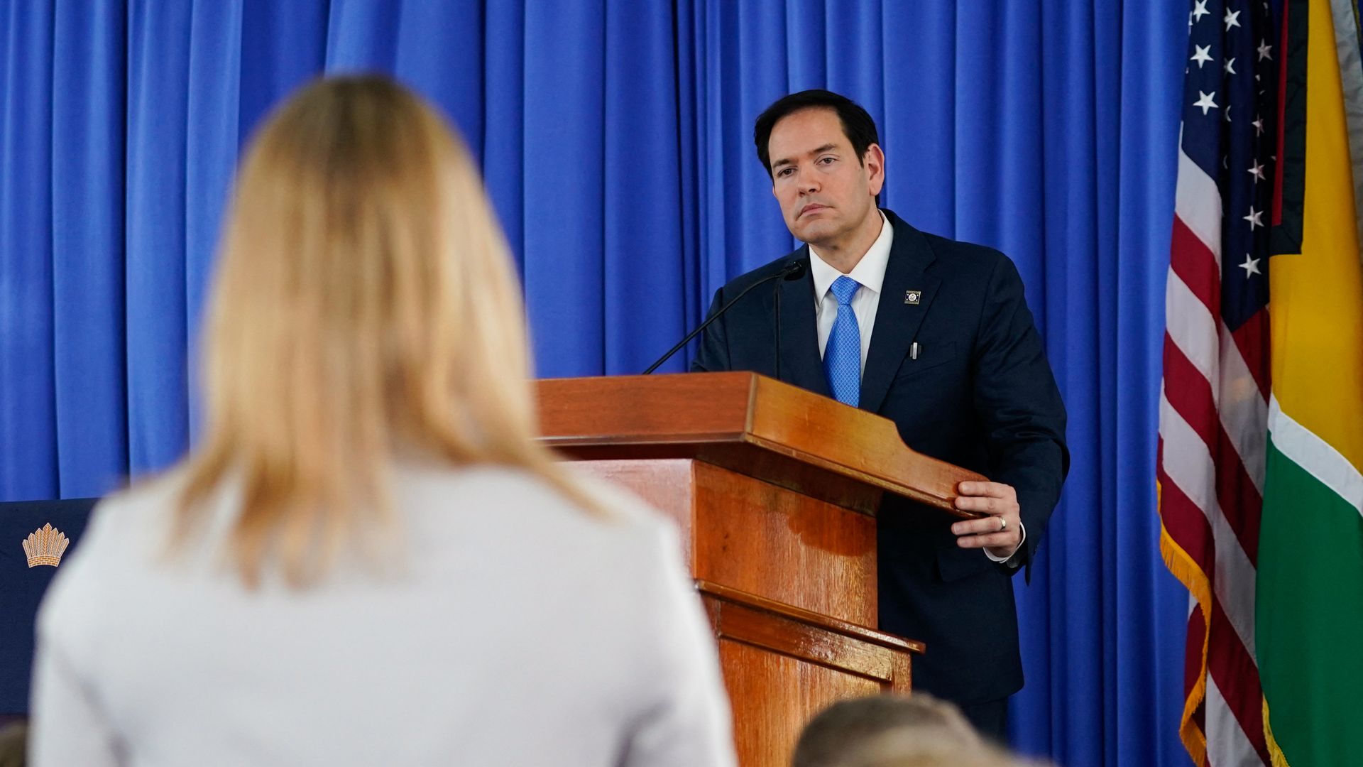 Marco Rubio listens from behind a podium as a reporter asks a question.