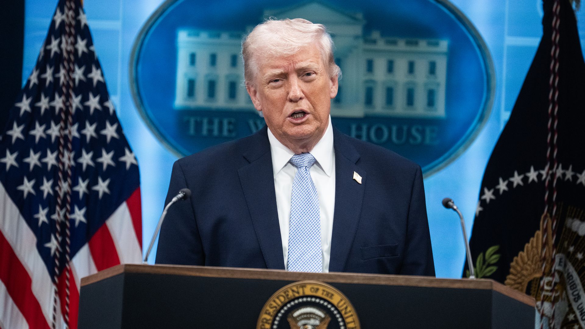 A man with light hair in a dark suit and light blue tie speaks at a podium bearing the presidential seal. A blue backdrop with a White House emblem and American flags flanks him.